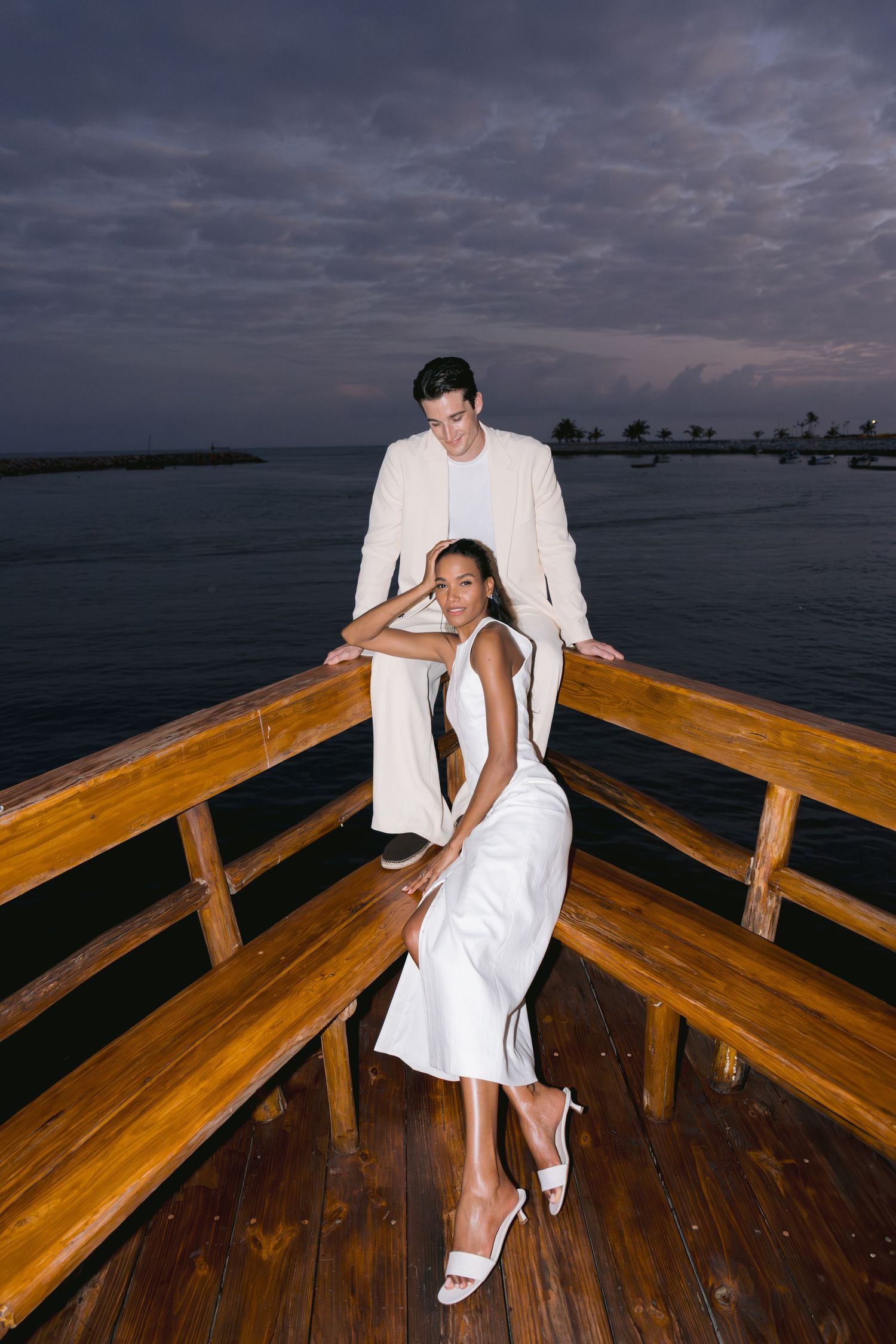 Romantic couple in white outfits share intimate moments on a wooden pier at sunset overlooking the water.