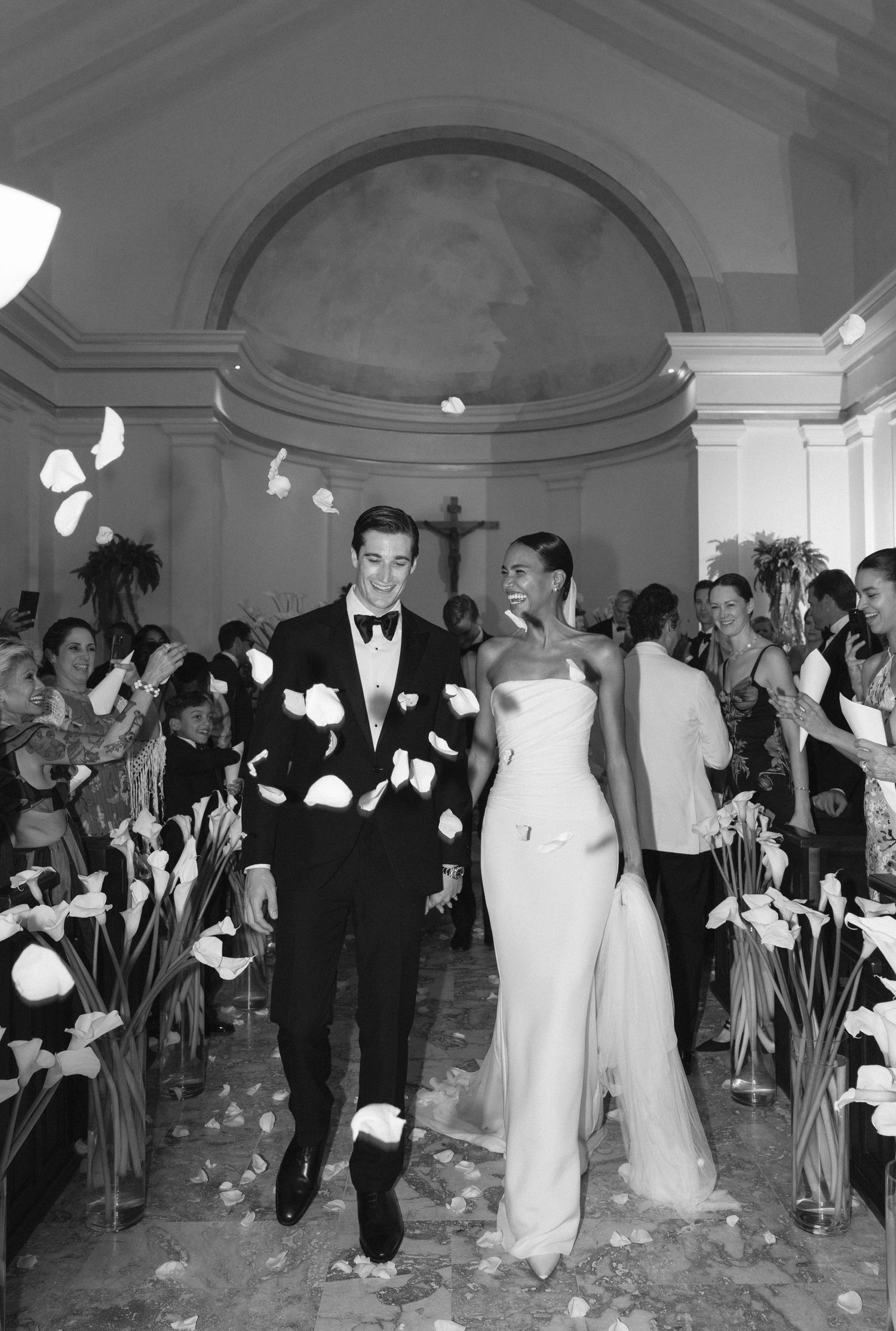 A wedding couple exits their ceremony while guests shower them with flower petals in an elegant domed venue.