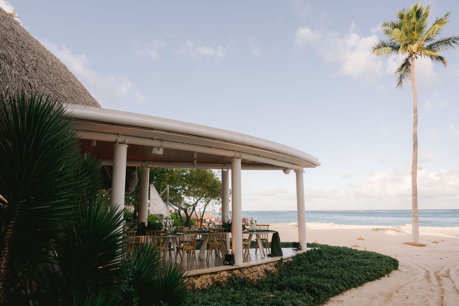 A beachside restaurant pavilion with thatched roof and white pillars overlooking a tropical beach with palm trees at sunset.