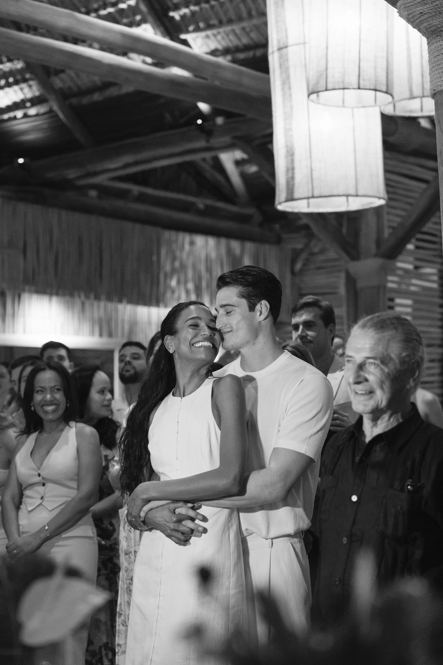 Black and white photograph capturing a tender romantic moment at a celebration with guests smiling in the background.