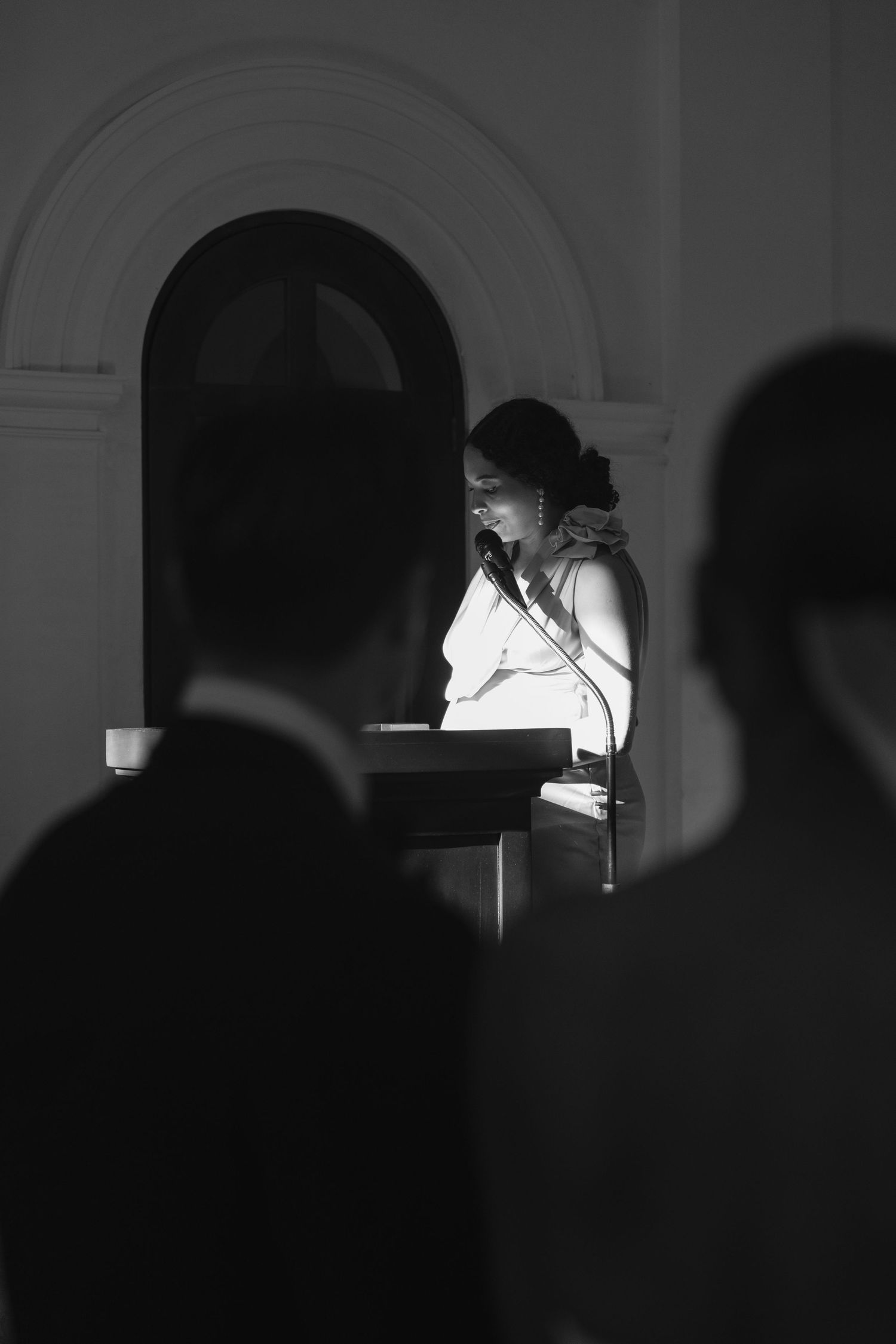 A speaker in a white top stands illuminated at a podium while dark silhouettes of audience members are visible in foreground.