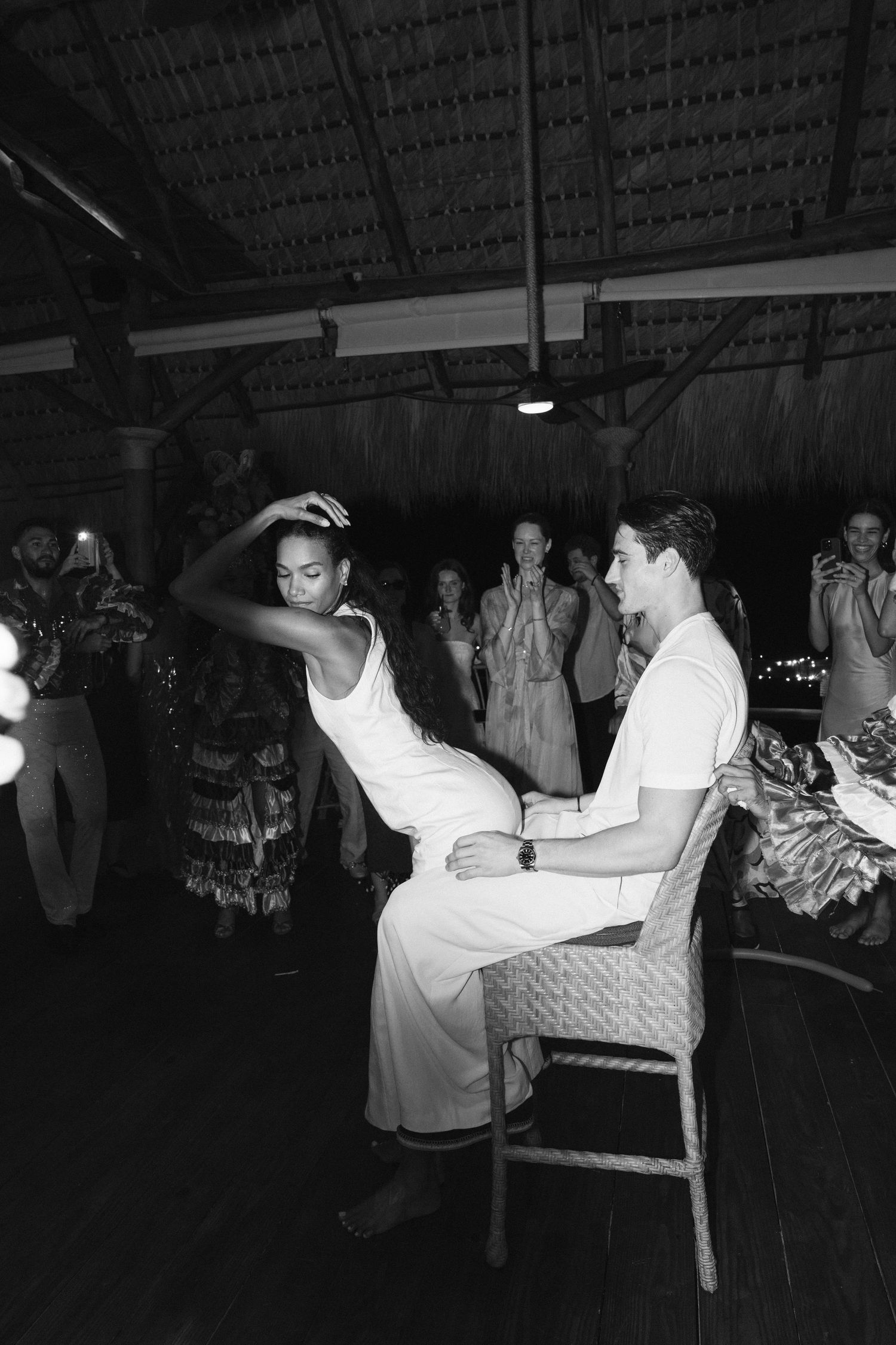 A couple dances together at a social event, with one person seated in a chair and the other moving energetically.