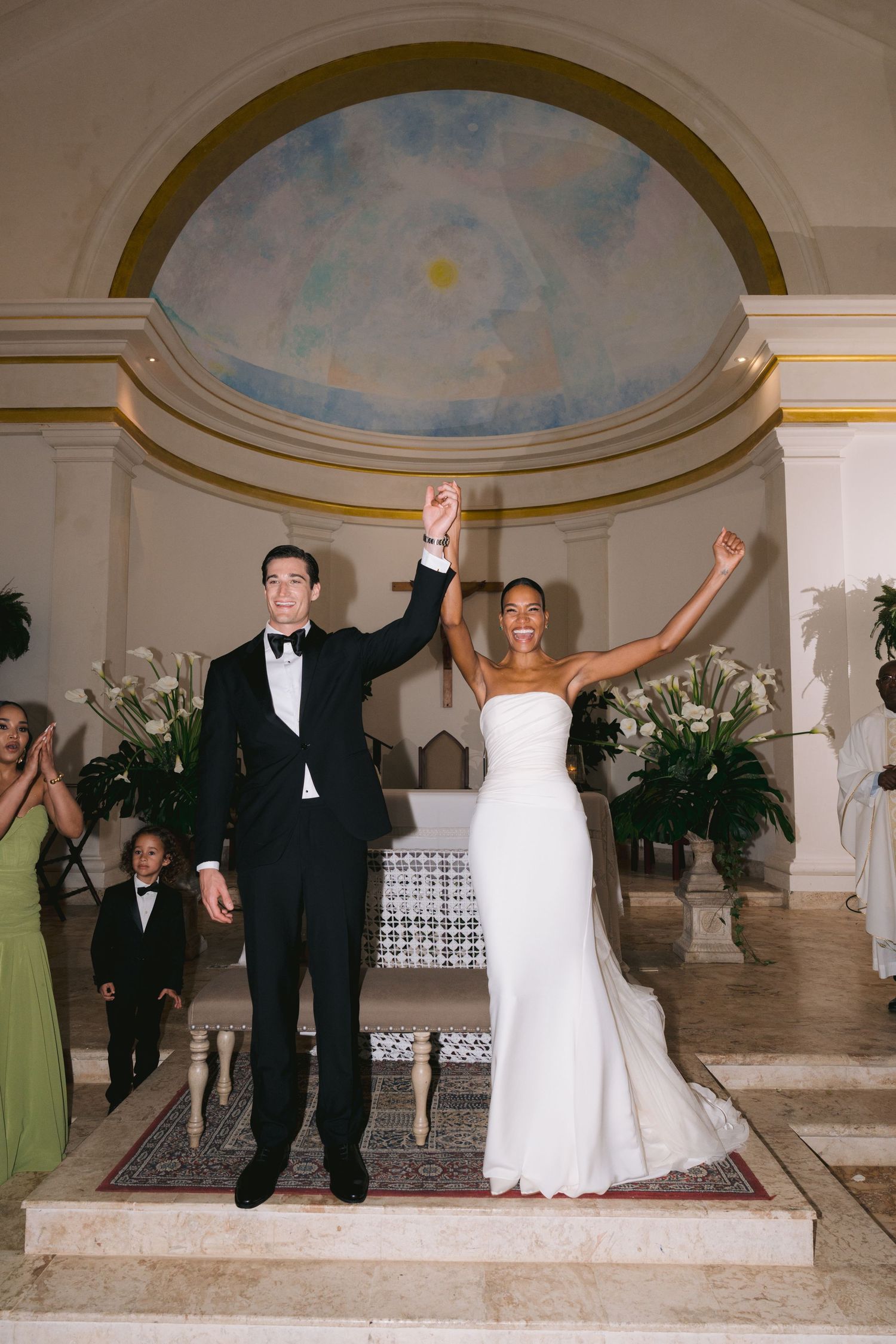 A couple in formal wedding attire celebrates with coordinated playful jumping poses inside an elegant marble ballroom.