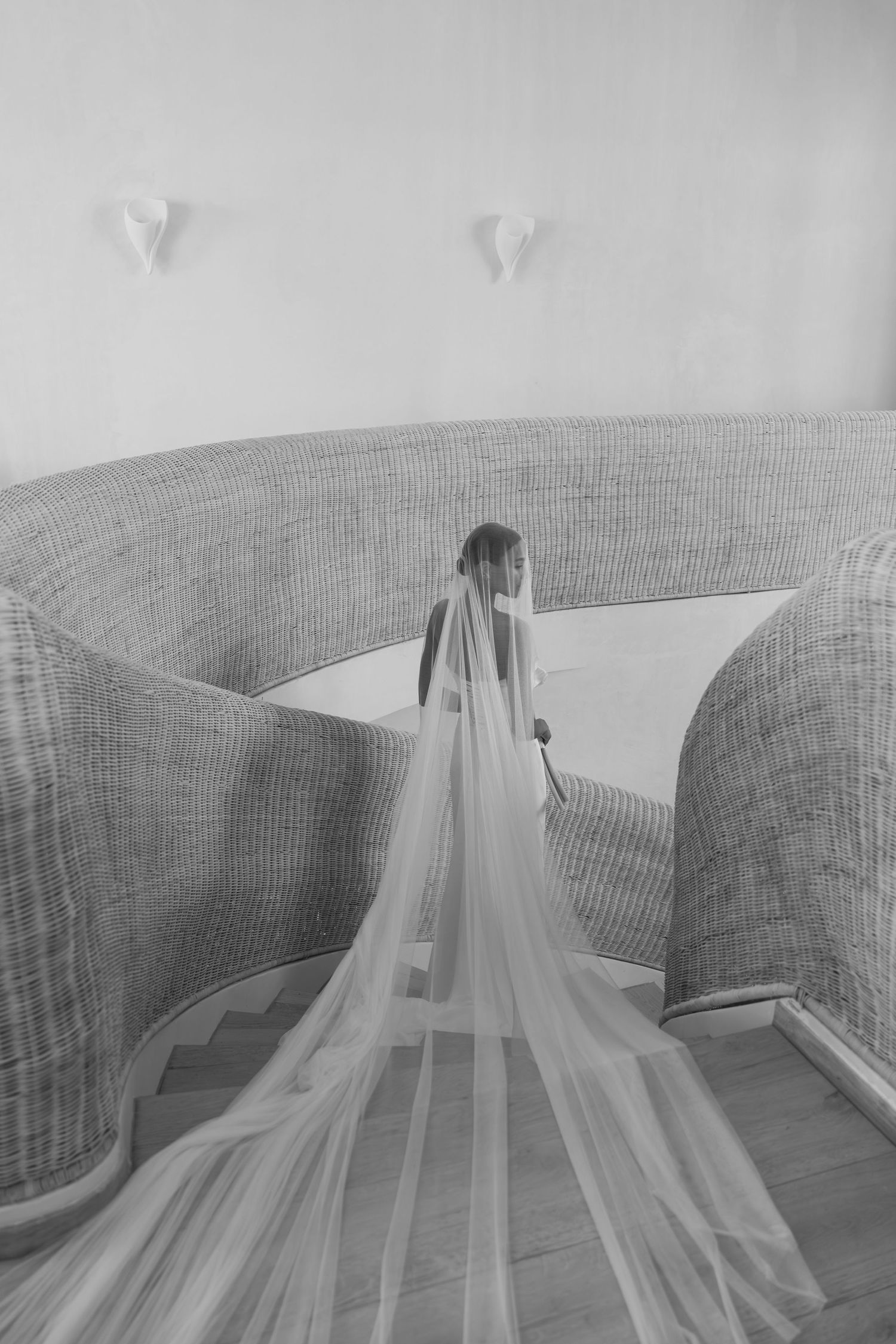 Black and white photograph of a bride in a long flowing veil standing between curved woven walls at a modern venue.