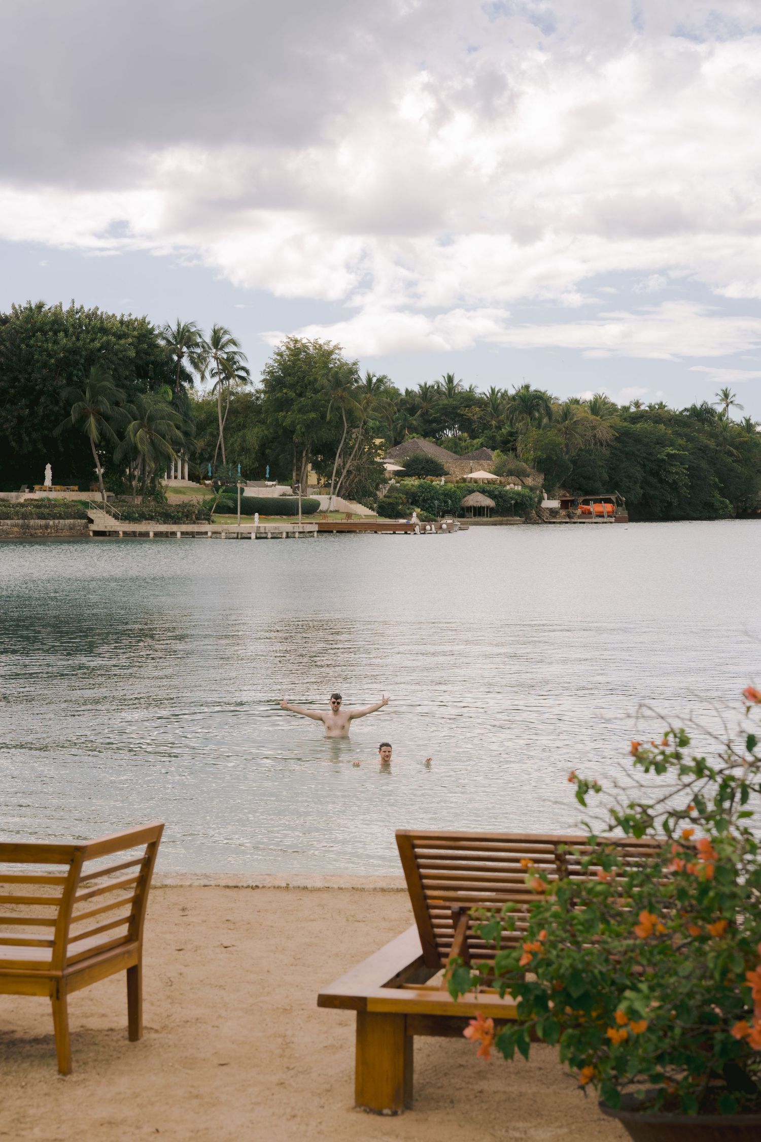 A series of wooden benches line a sandy beach overlooking a tranquil lagoon with palm trees and cloudy skies in Hawaii.