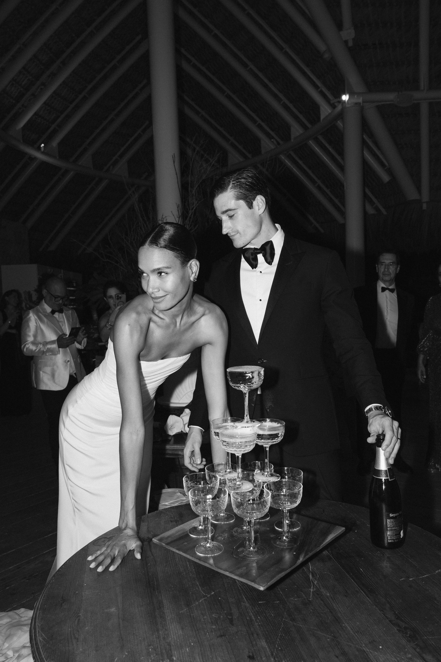 A black and white photo of a couple at an elegant event pouring champagne into stacked coupe glasses on a table.