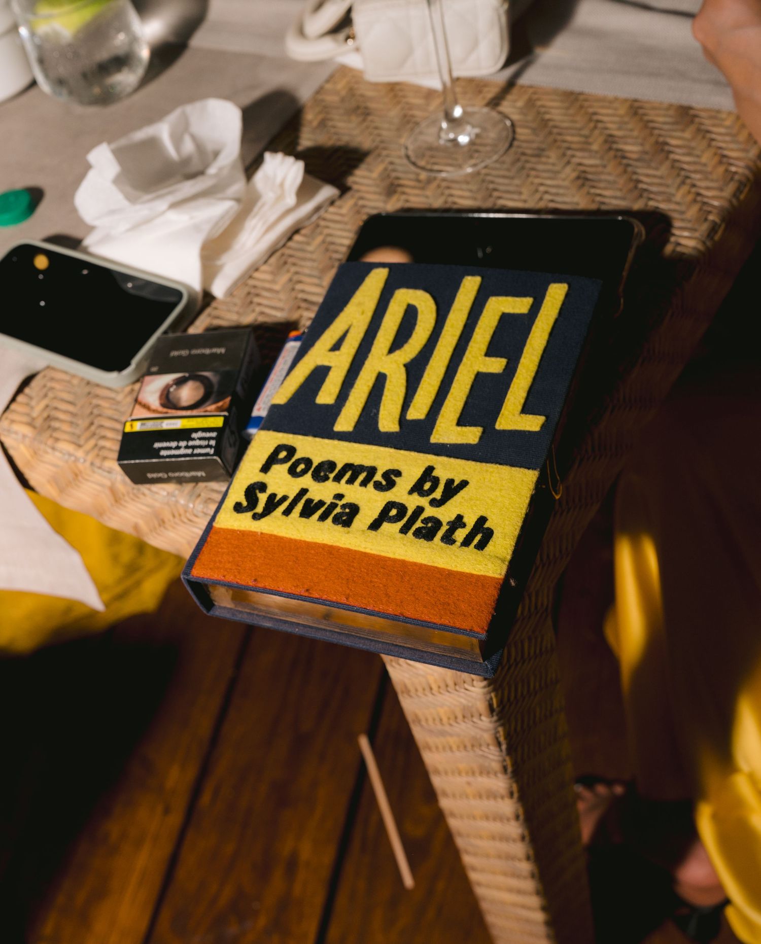 A vintage copy of 'Ariel' poetry book by Sylvia Plath sits on a wooden surface next to a wine glass.