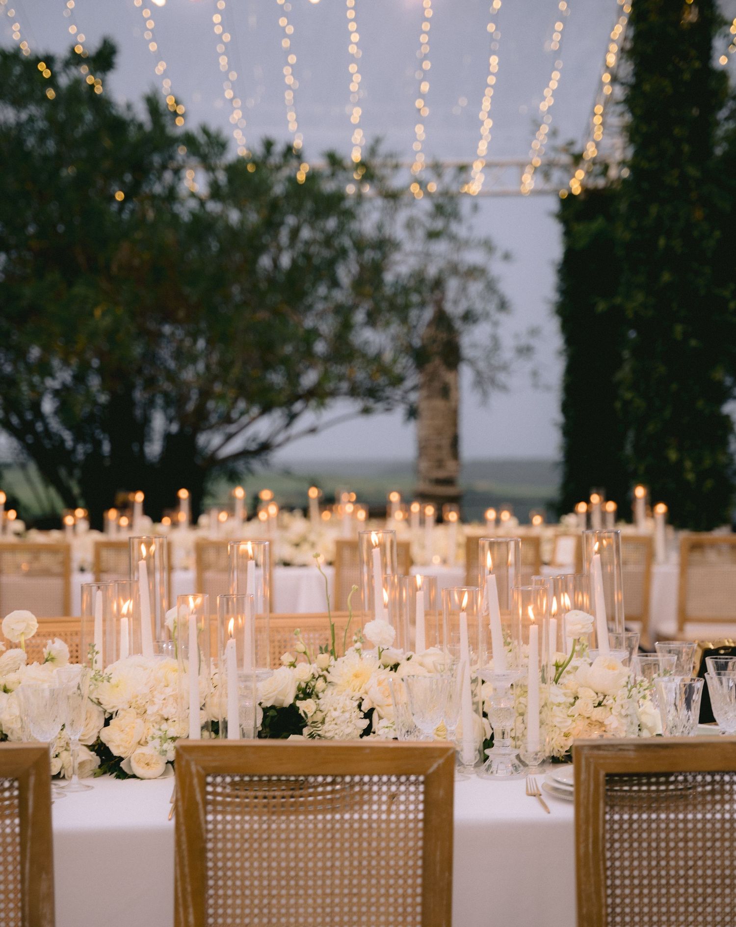 Elegant wedding reception table with glass candle holders and white floral centerpieces under string lights.