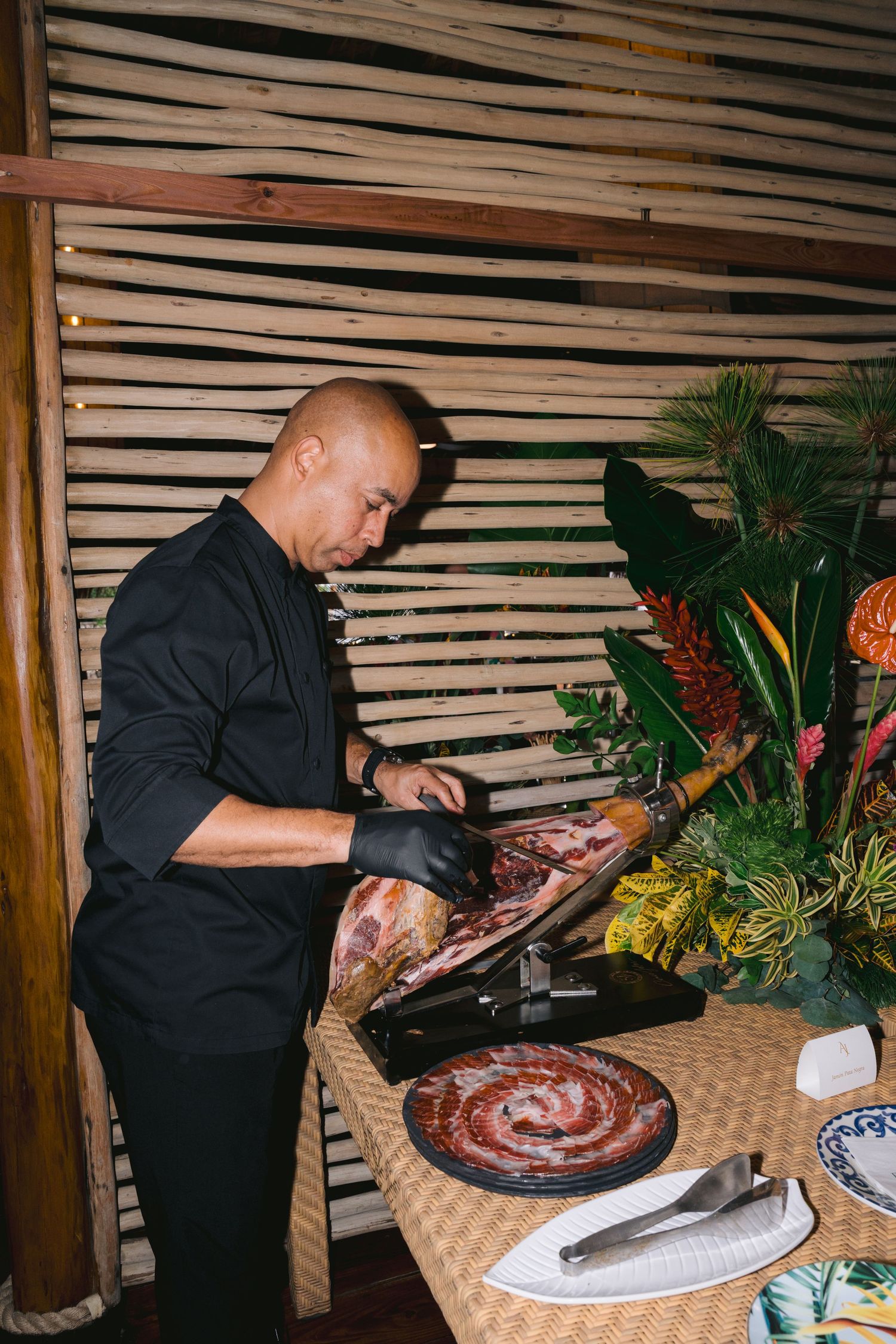 Chef in black uniform prepares meat at rustic table with woven surface and bamboo wall decor.