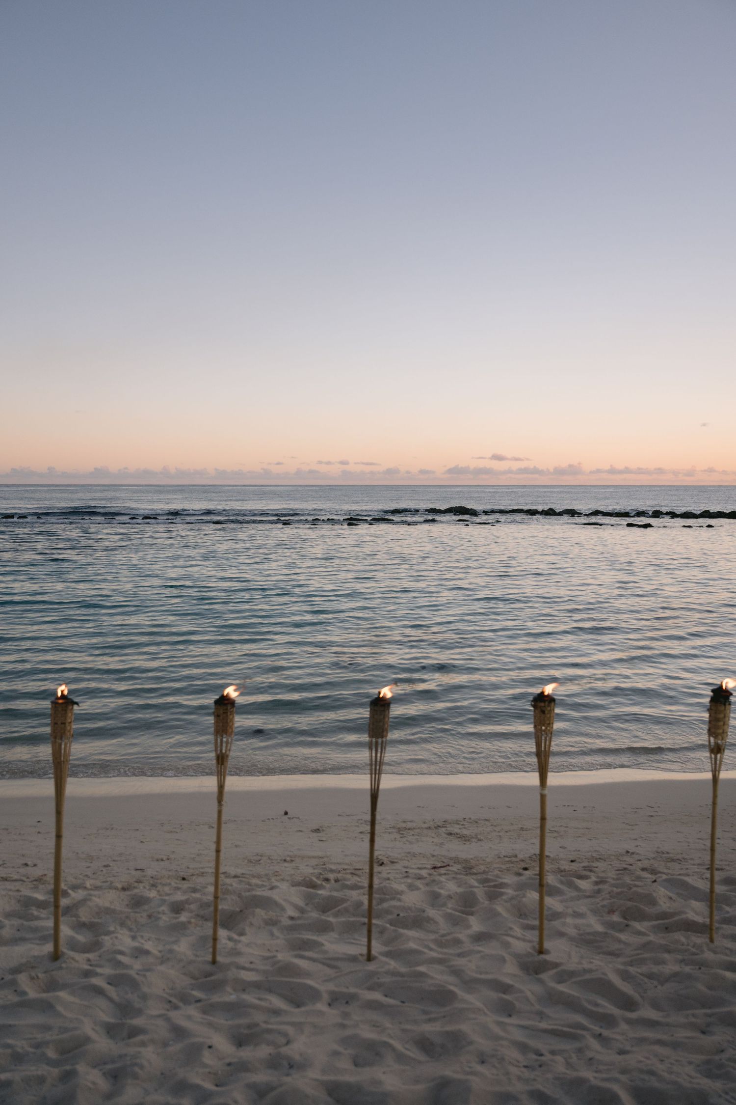 Tiki torches line a sandy beach at sunset with gentle waves rolling onto the shore and a soft pink sky overhead.