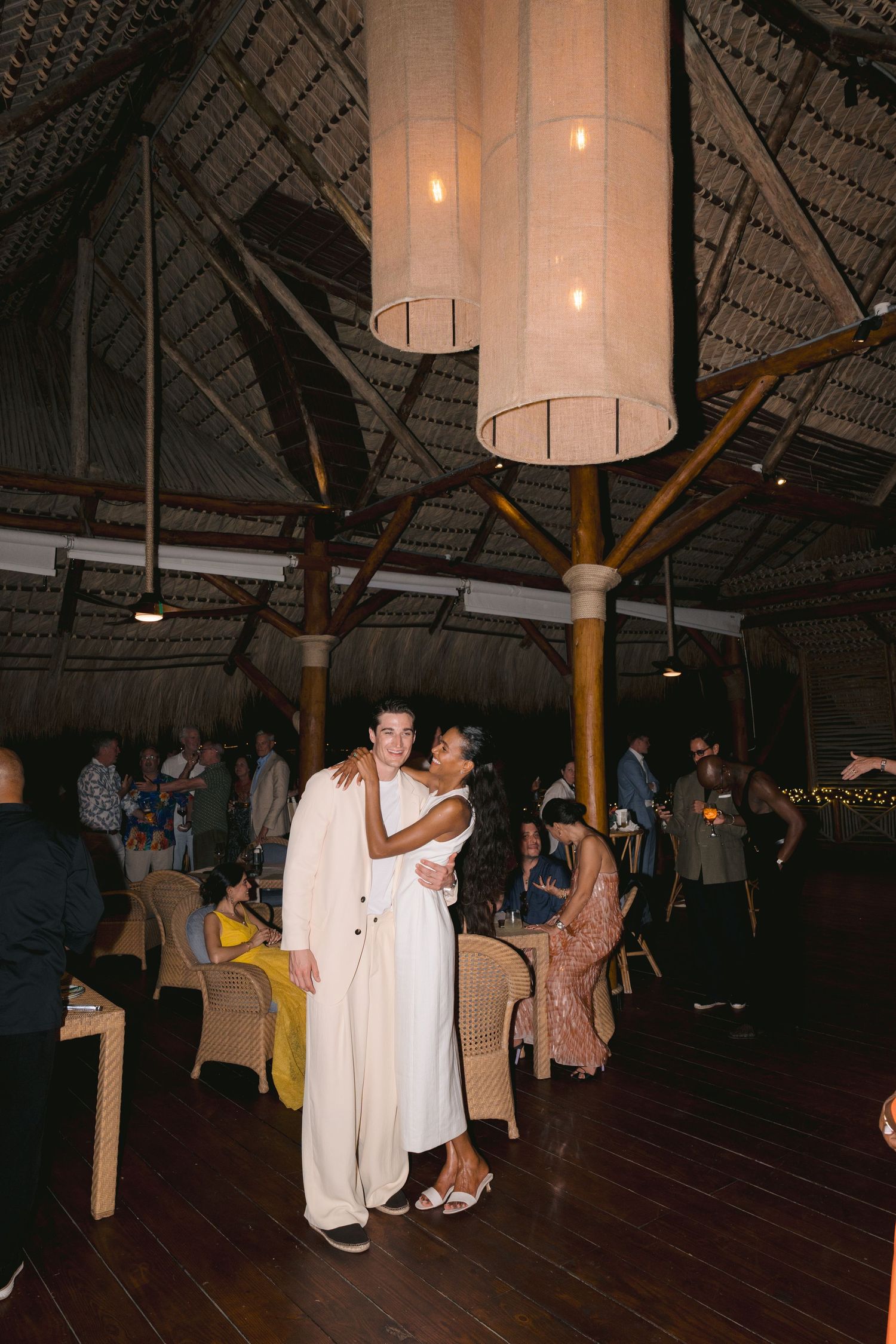 Couple in white outfits dancing at a tropical reception venue with bamboo pillars and cylinder pendant lights.
