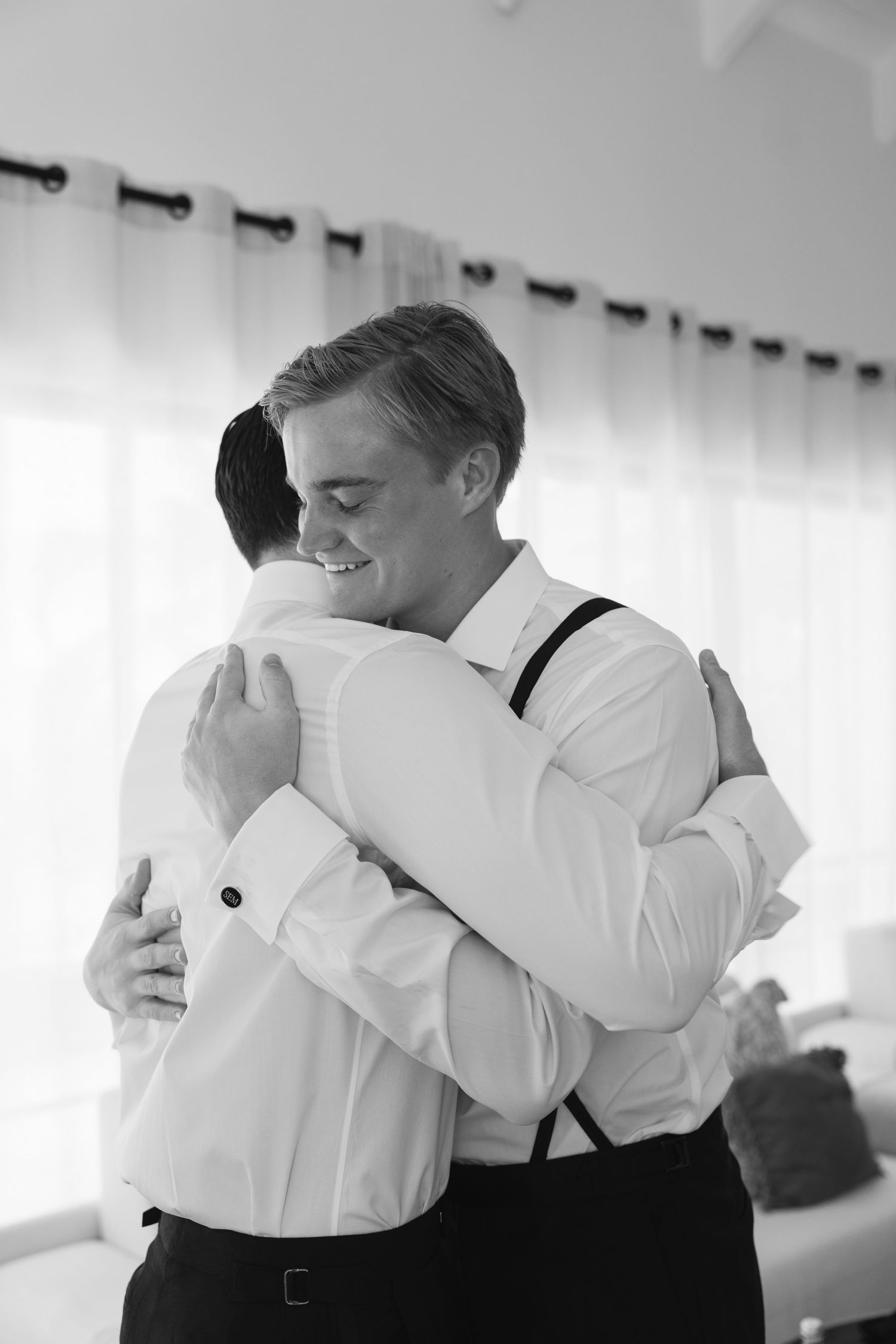 Two people share a tender embrace while wearing formal attire with white shirts and suspenders in black and white.