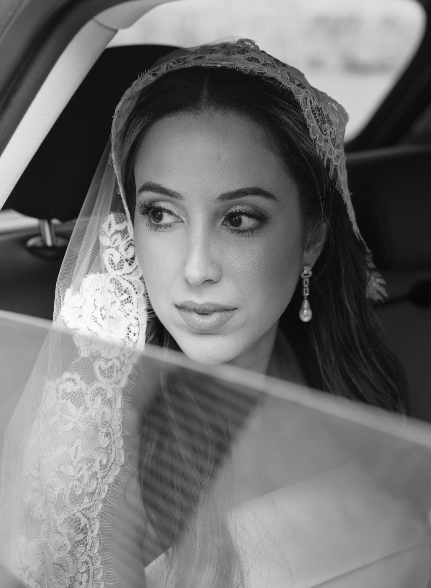Black and white portrait of a bride in elegant lace veil and sparkling earrings inside a car.