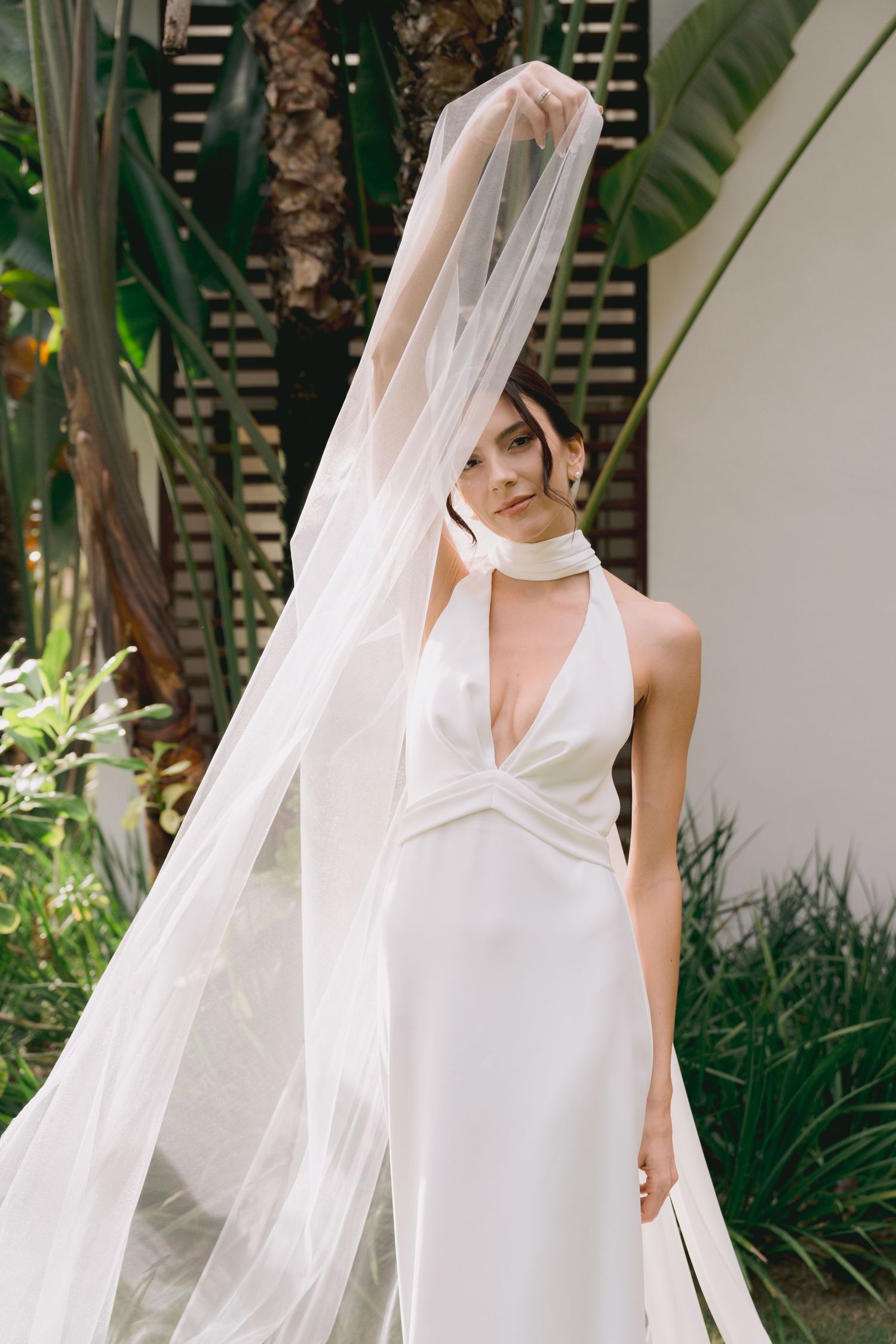 Elegant white halter wedding dress with flowing veil against a tropical garden backdrop.
