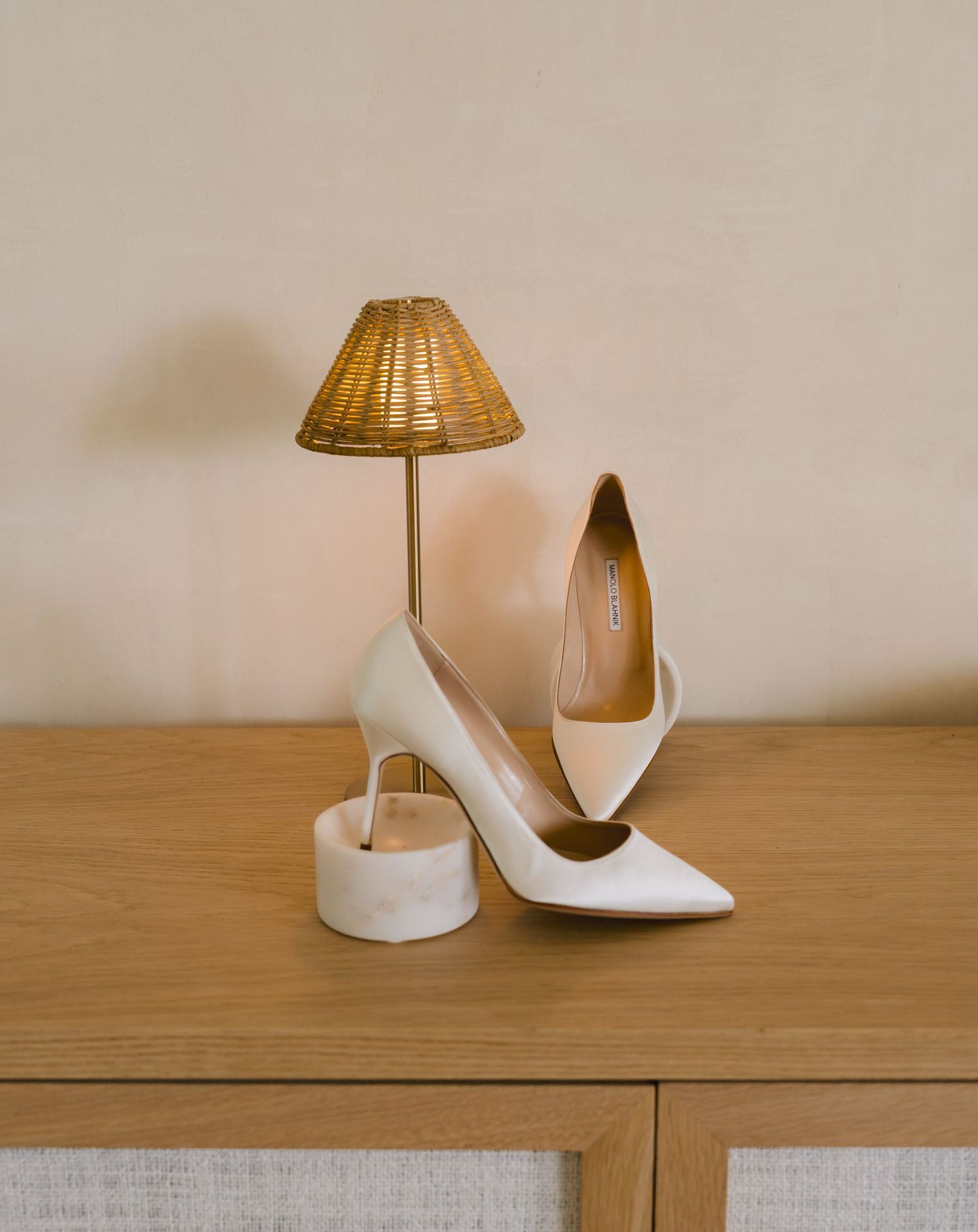 Elegant white stiletto heels and a small table lamp with a woven shade displayed on a wooden surface.