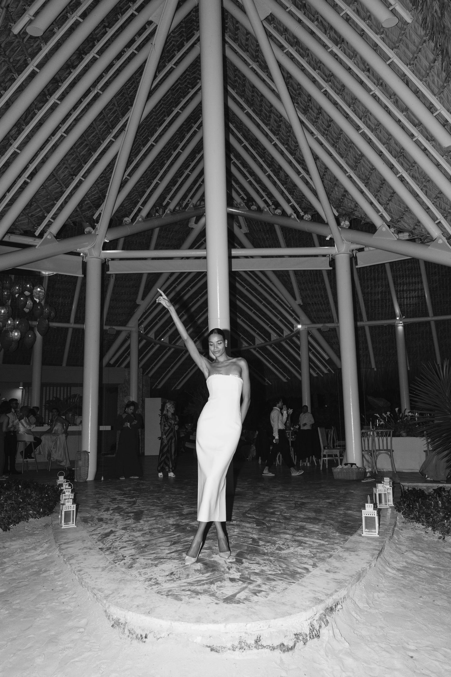 A black and white photograph of an elegant figure in a white dress standing under a geometric gazebo structure at night.