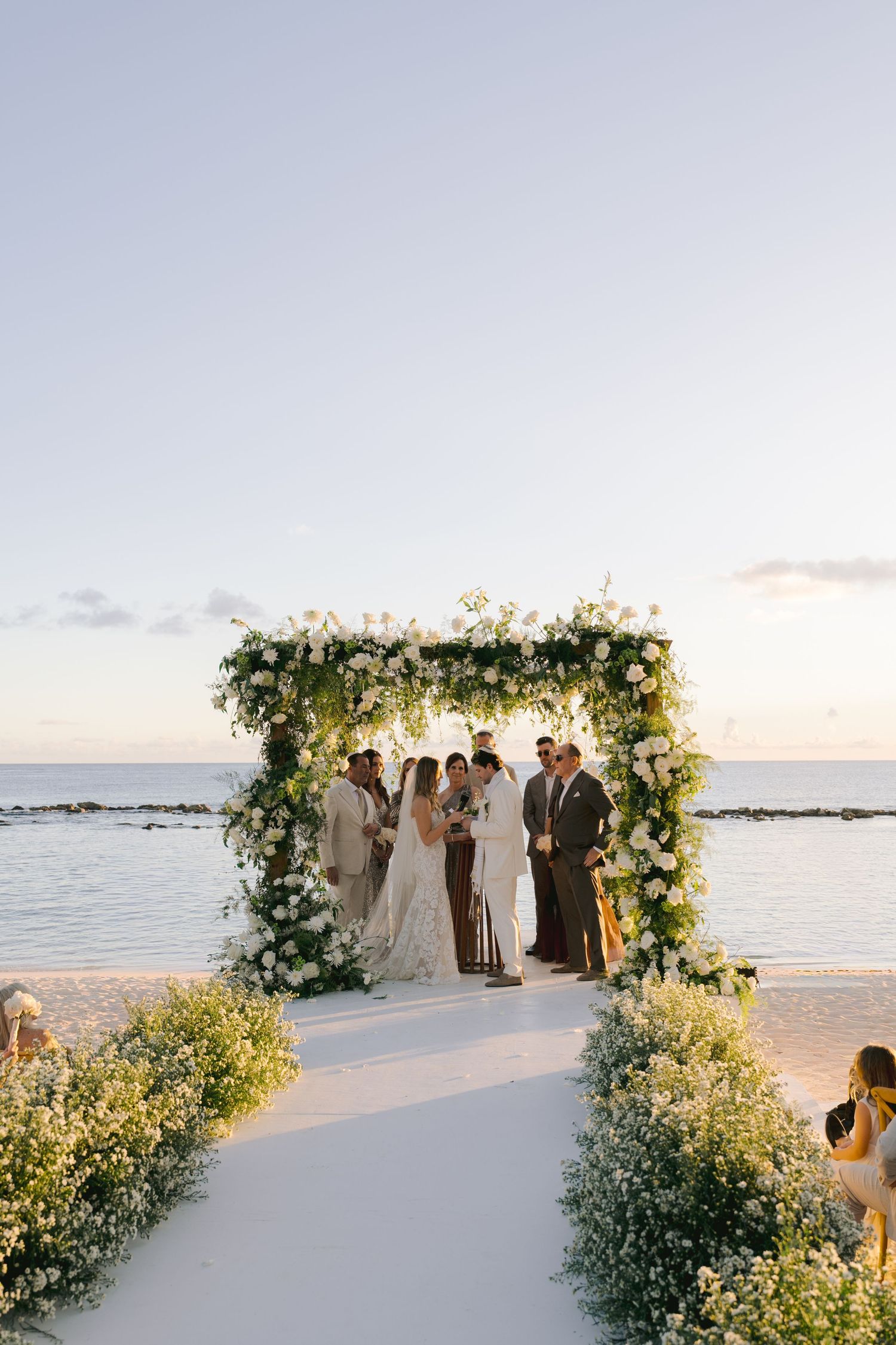 A beachside wedding ceremony setup with a floral arch and chairs arranged on white sand at sunset.