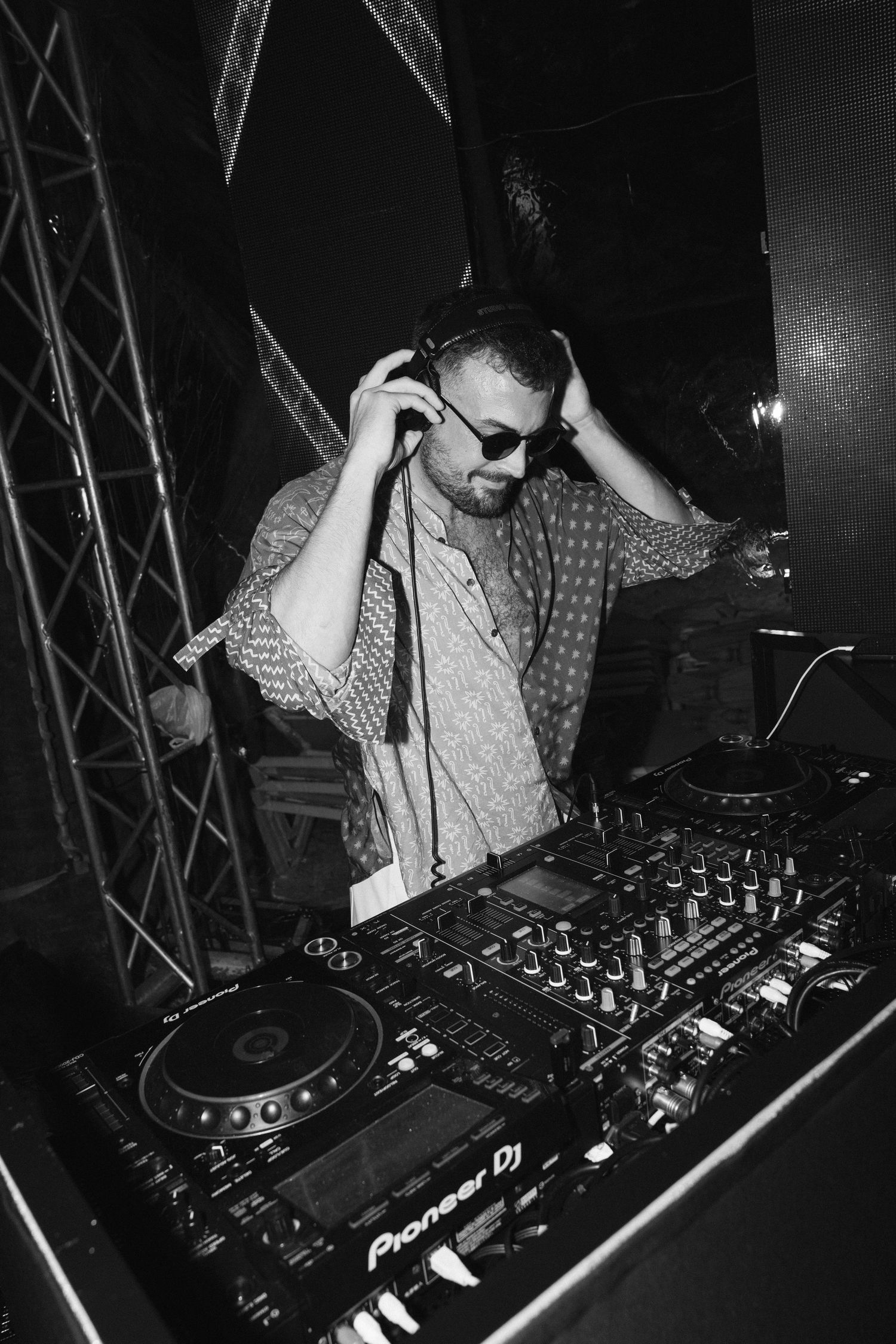 A DJ performs at a nightclub behind Pioneer decks wearing sunglasses and adjusting headphones in black and white.