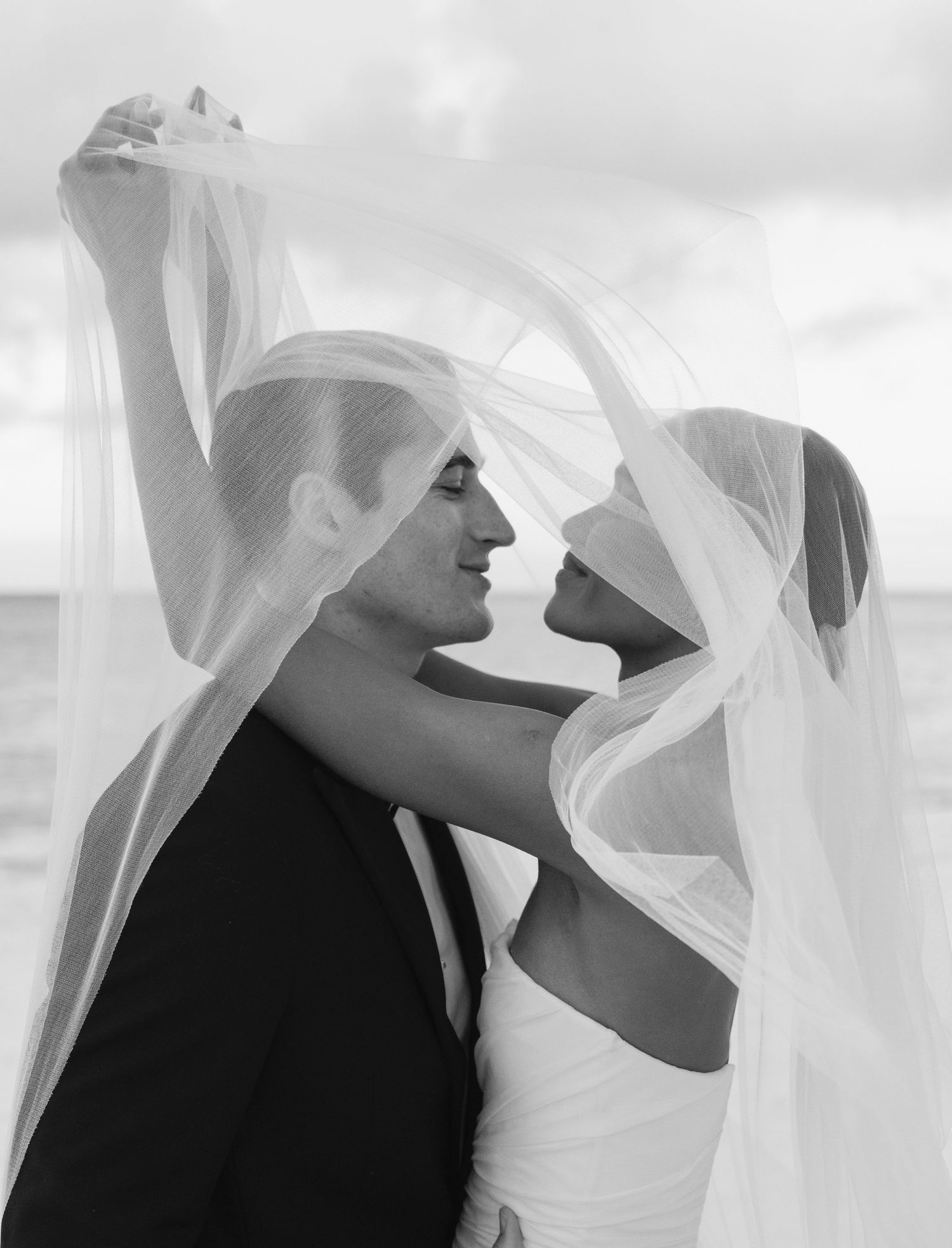 A black and white wedding photograph showing a couple in formal attire with flowing veils caught in a romantic breeze.