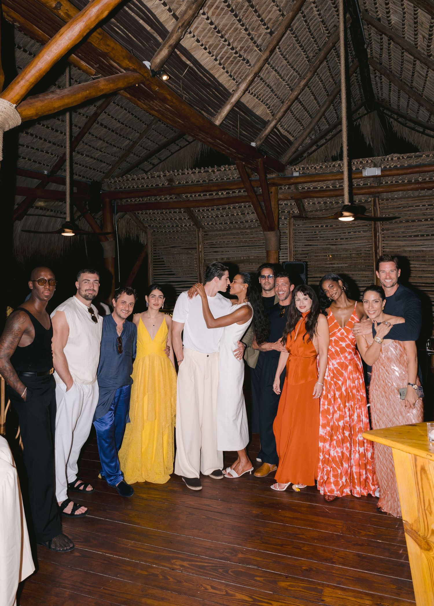 A wedding celebration with guests in colorful formal attire gathered inside a rustic wooden barn with exposed beams and lighting.