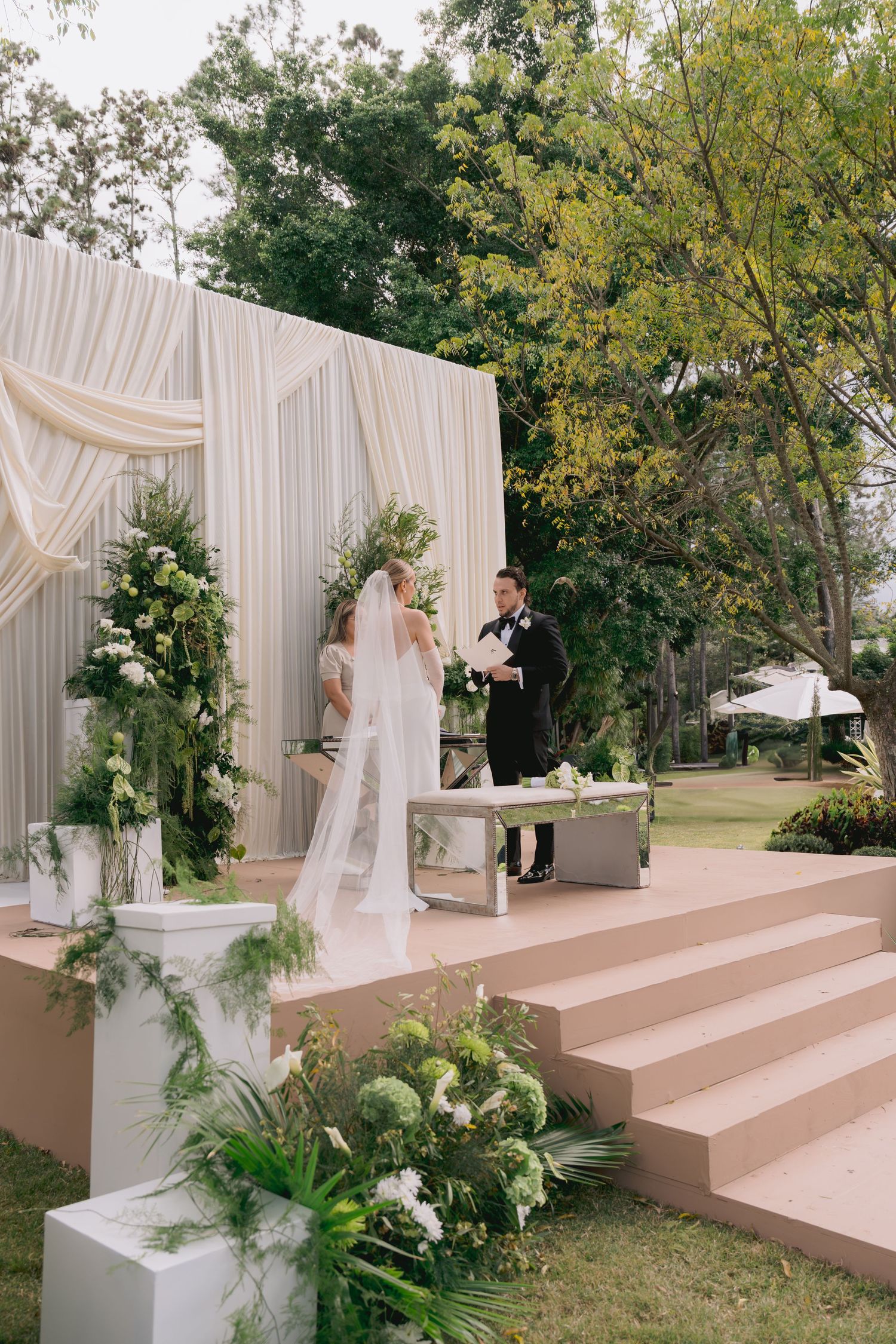 Outdoor wedding ceremony setup with white draping and floral arrangements on elevated platform.