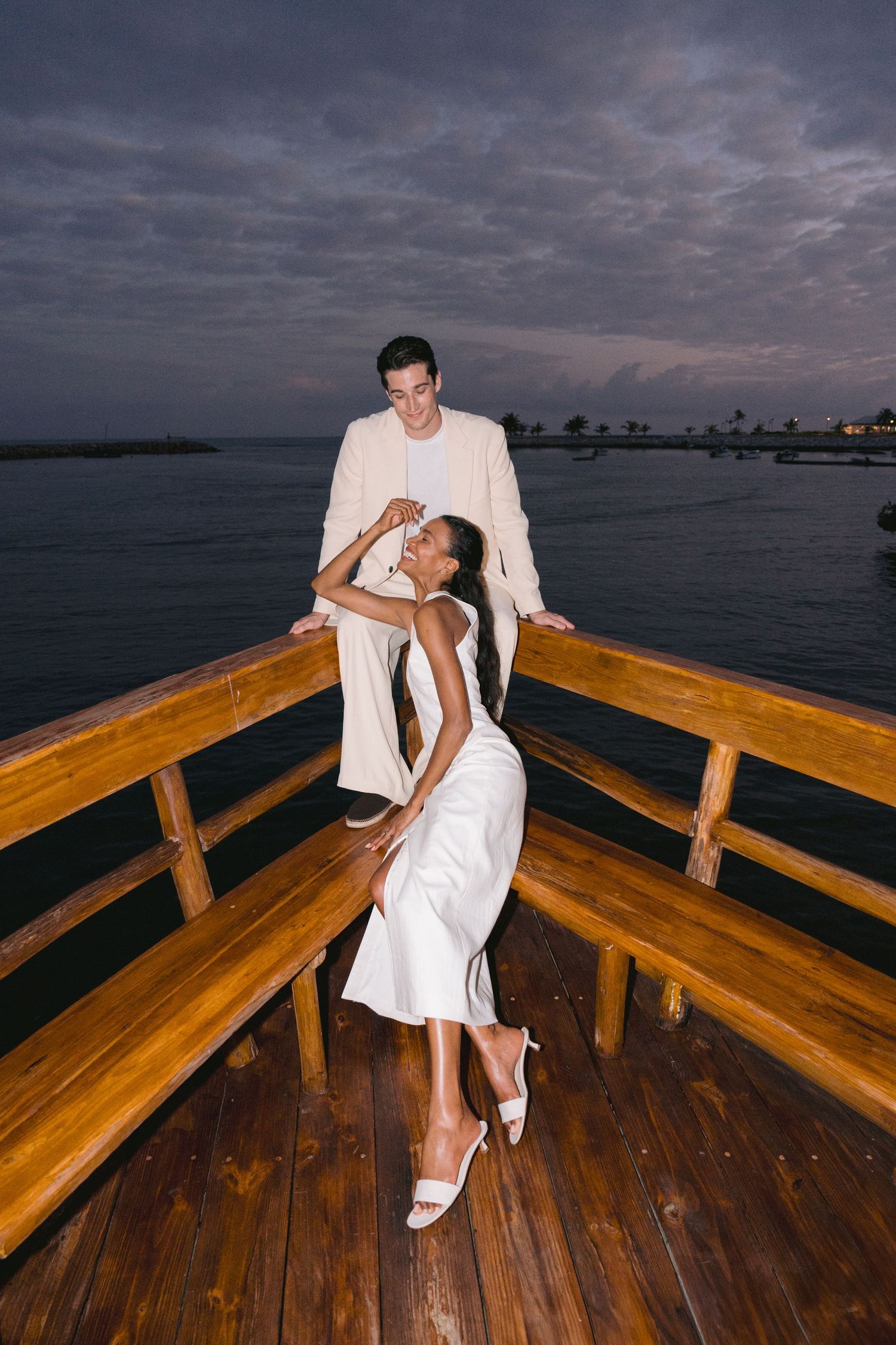 A couple in white formal attire strike a romantic pose on a wooden deck overlooking the ocean at dusk.