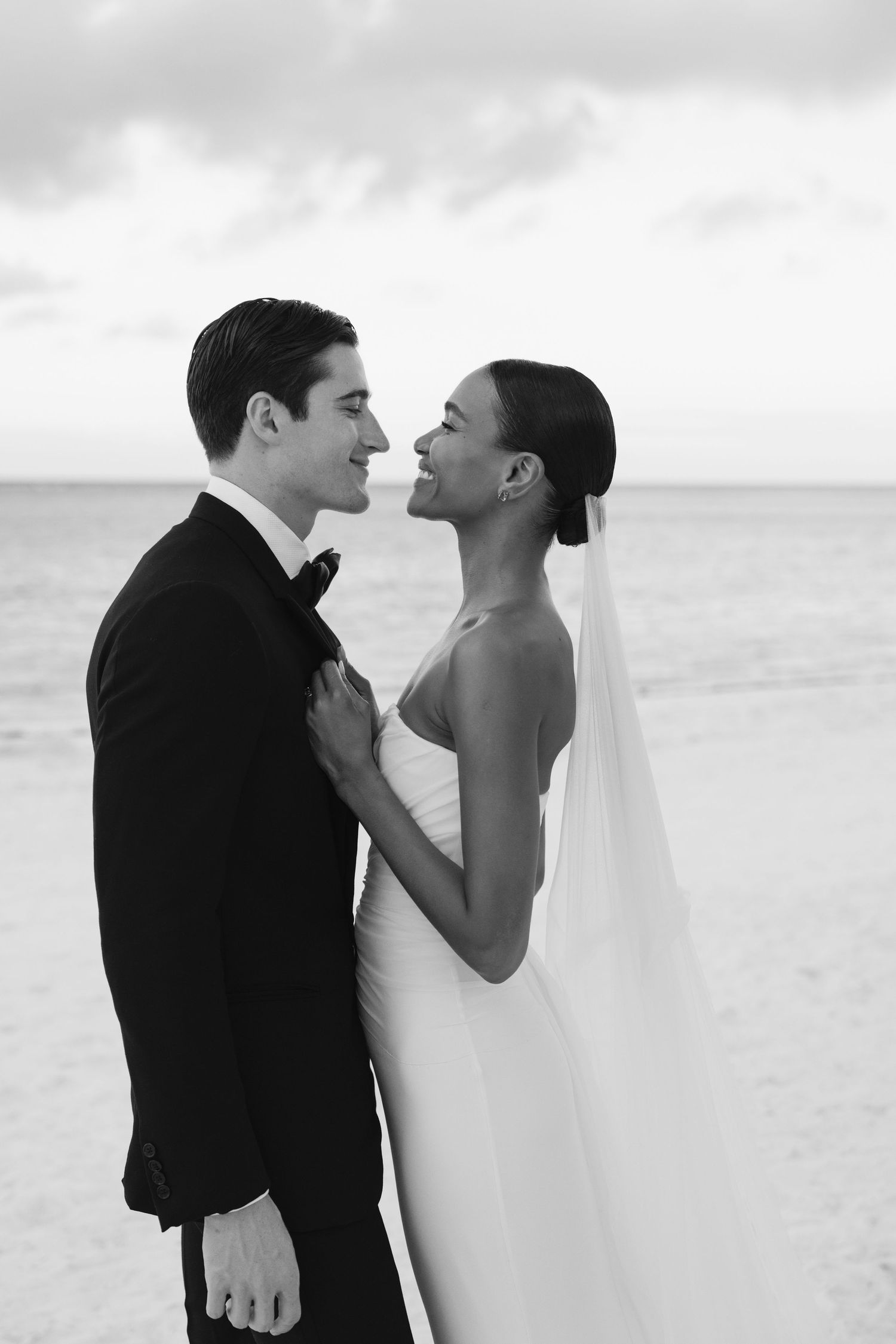 Black and white romantic beach wedding photo of a couple embracing by the ocean under cloudy skies.