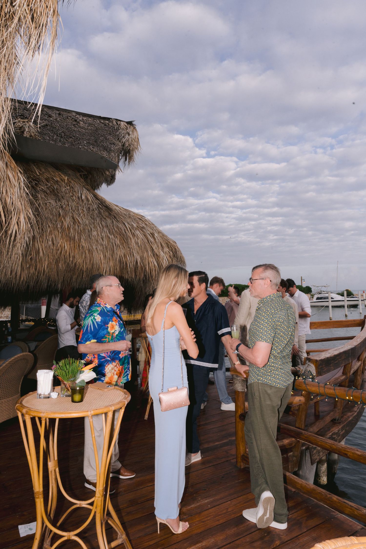 People gathered on a wooden deck under thatched roof palapas at a tropical waterfront bar or restaurant.