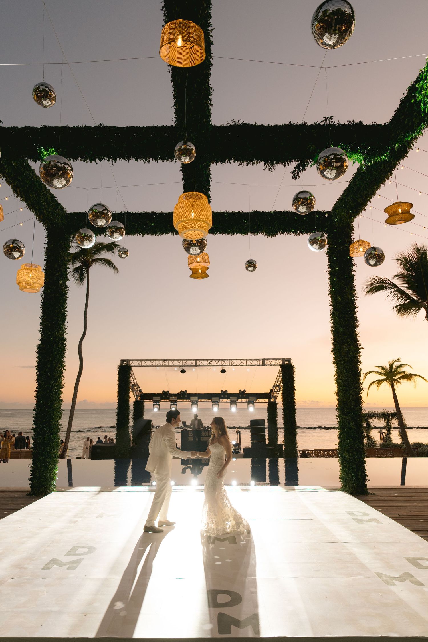 Elegant beach wedding reception setup with white decor, hanging lights and palm trees silhouetted against sunset sky.