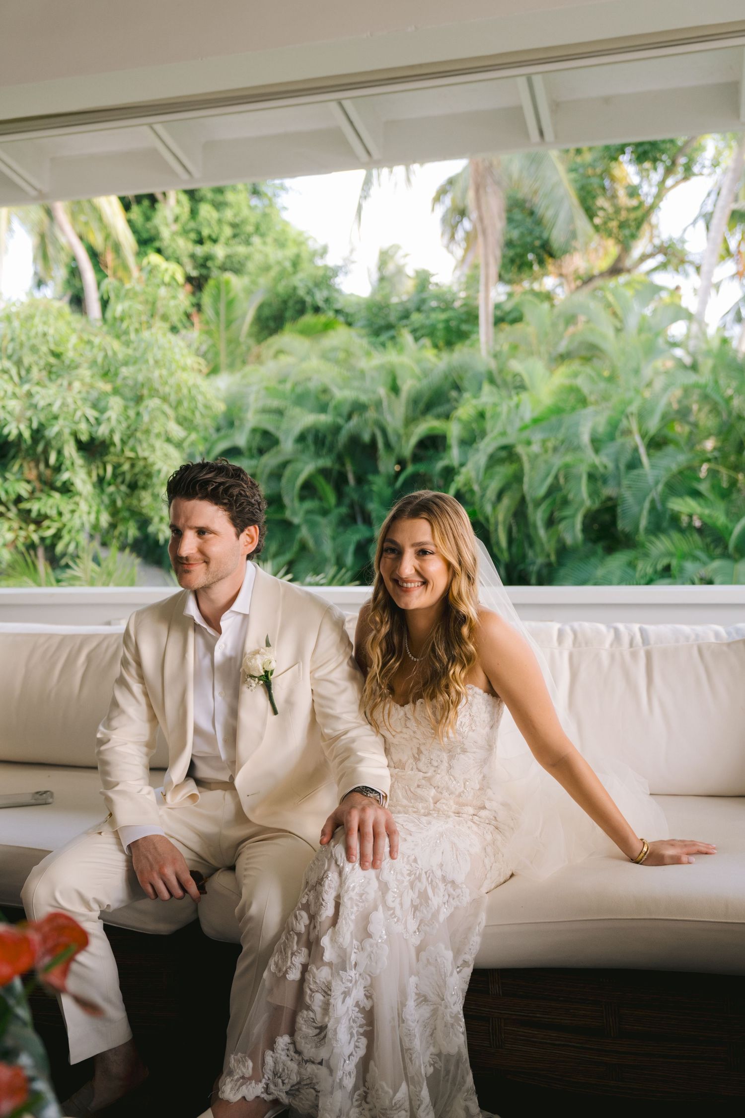 A bride and groom share a joyful moment while sitting together on a white couch surrounded by tropical greenery.