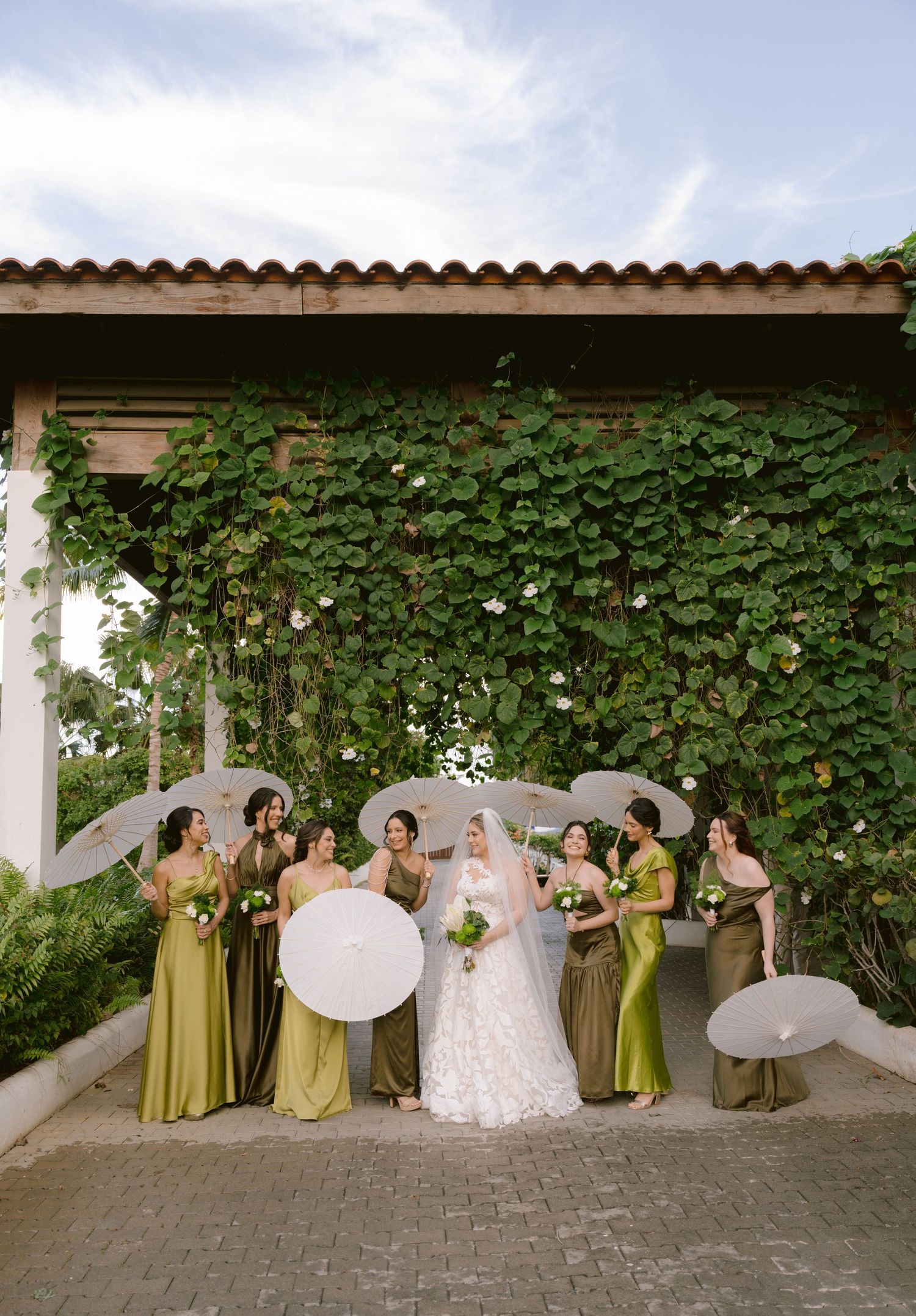 Bridal party with parasols poses against vine-covered wall wearing sage green dresses.