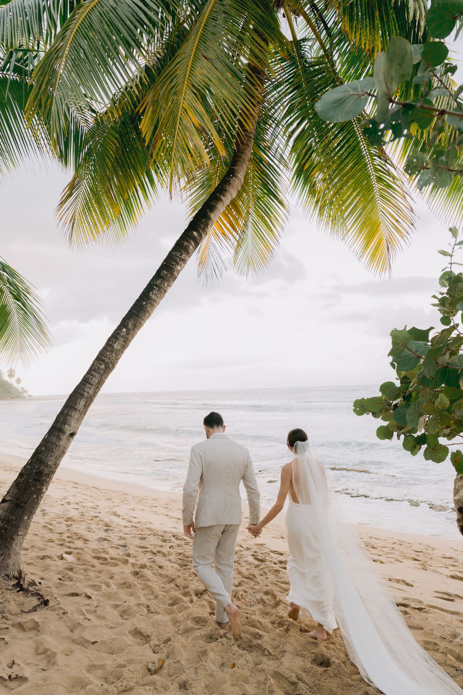 Newlyweds walking hand in hand on a tropical beach under swaying palm trees.