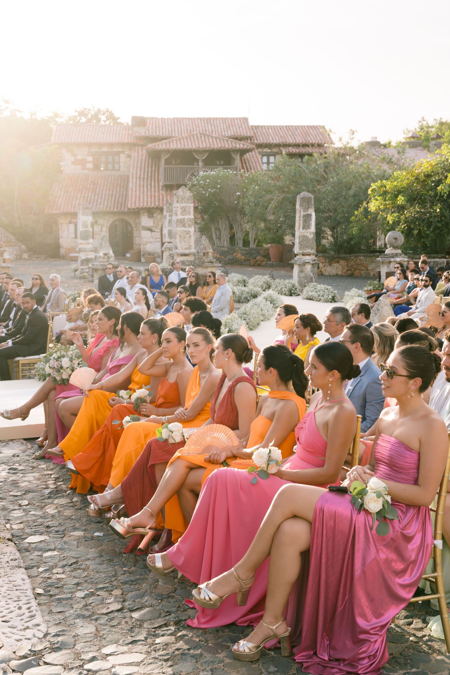 Wedding guests in colorful orange and pink dresses seated outdoors at rustic venue.