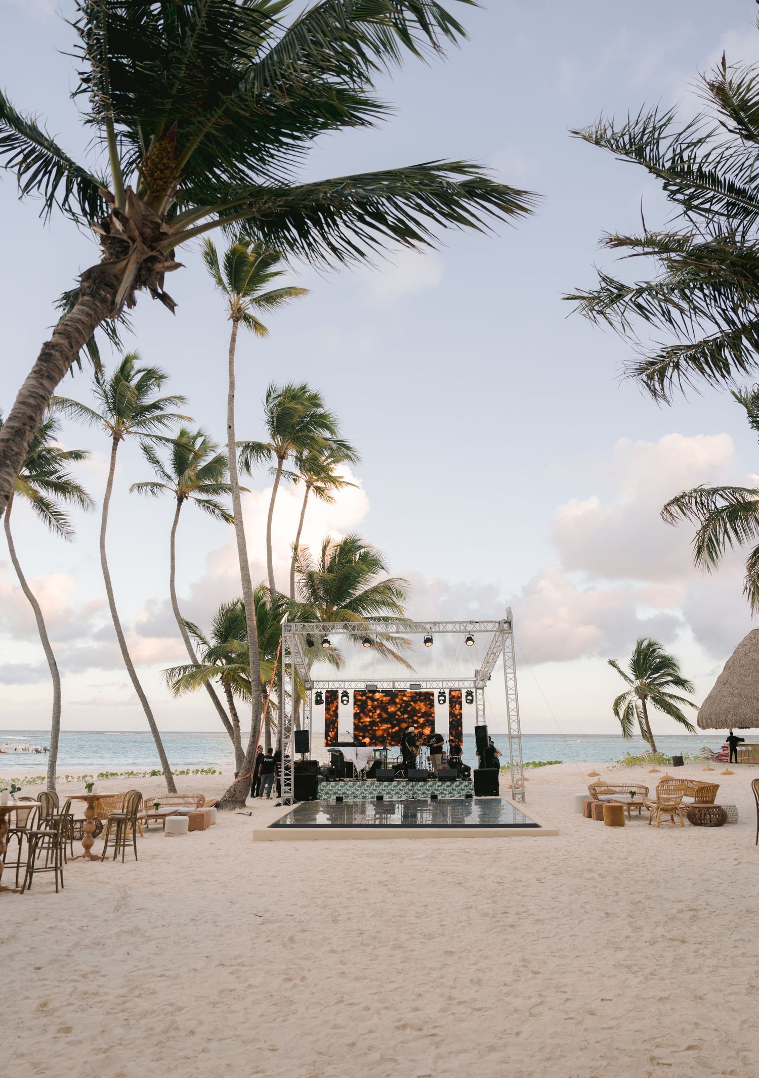 Tropical beach wedding setup with palm trees framing an elegant stage on white sand beside turquoise ocean waters.