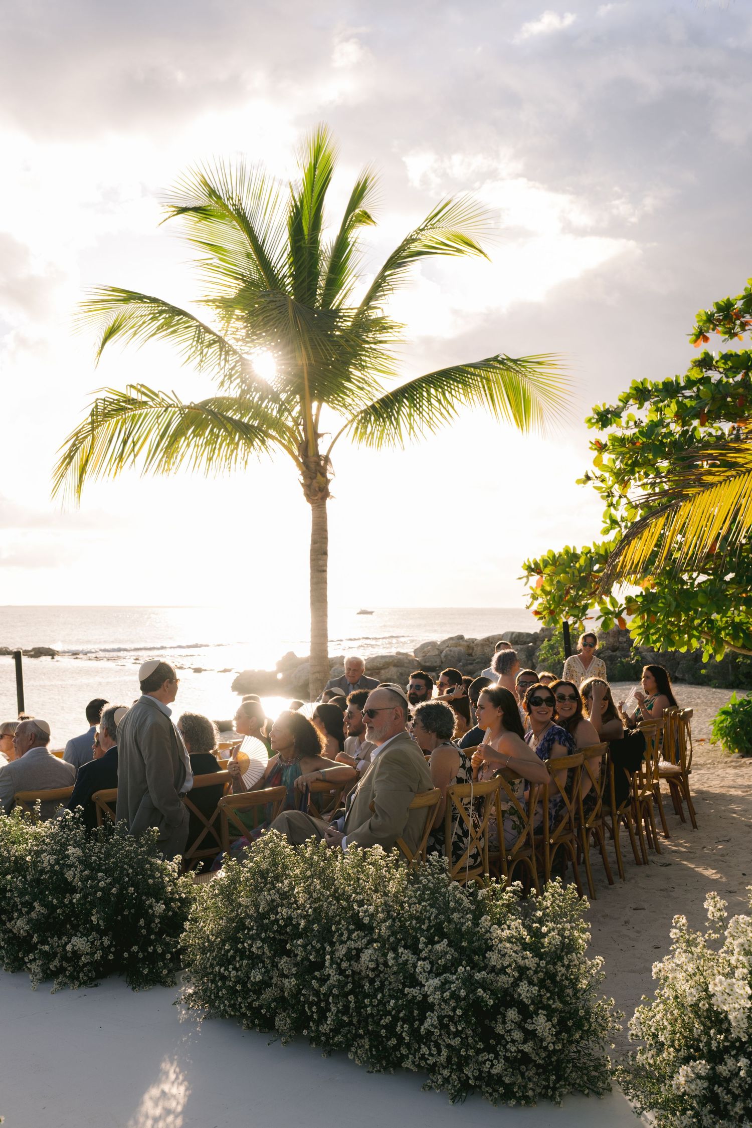 Wedding guests gather at a beachside ceremony under palm trees during sunset in a tropical destination.