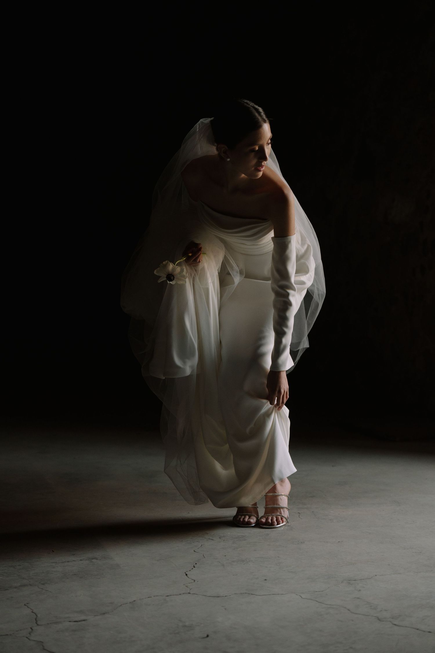 A dramatic low-light portrait of a bride adjusting her wedding dress.
