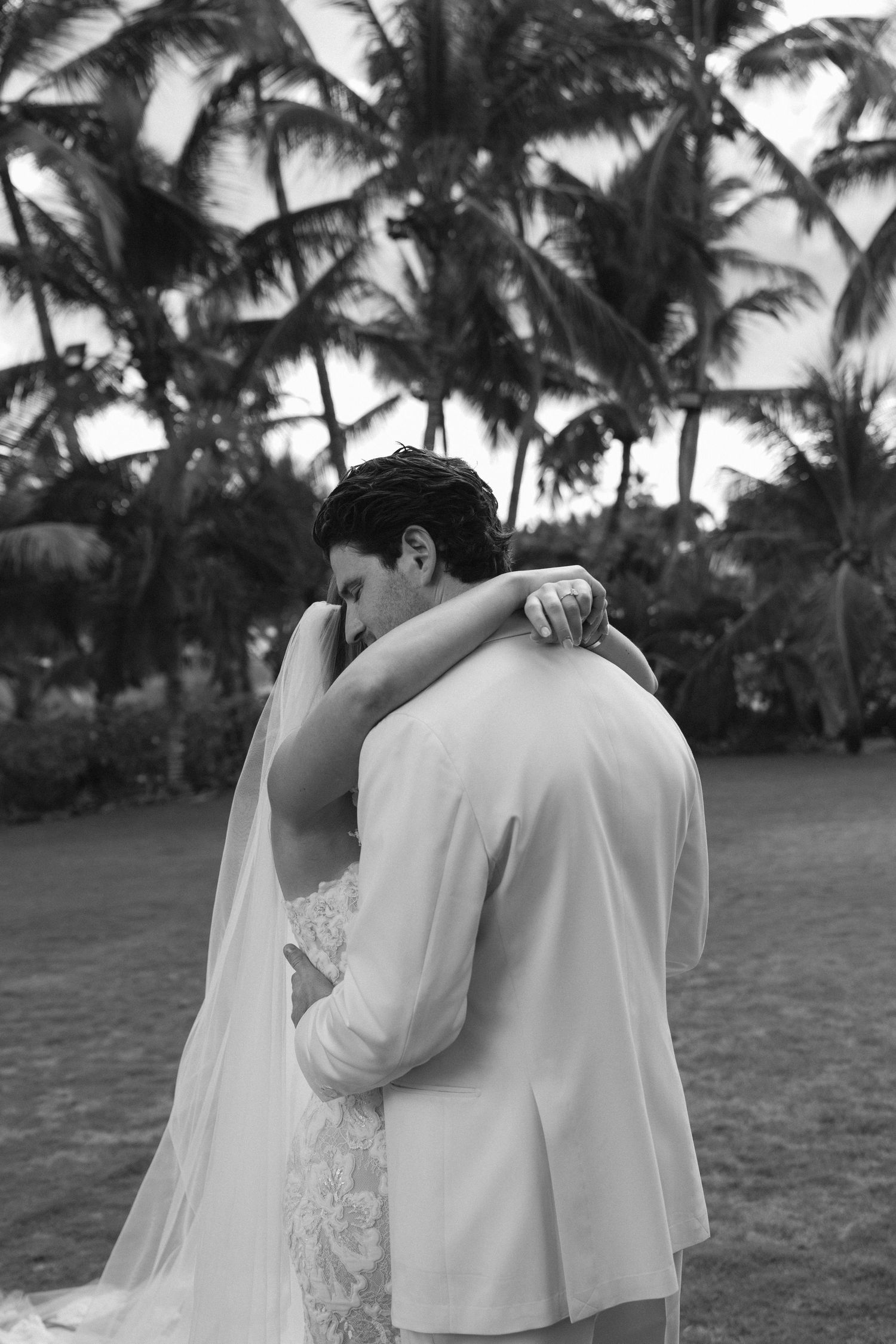 A couple embraces during their beach wedding ceremony surrounded by palm trees at sunset.