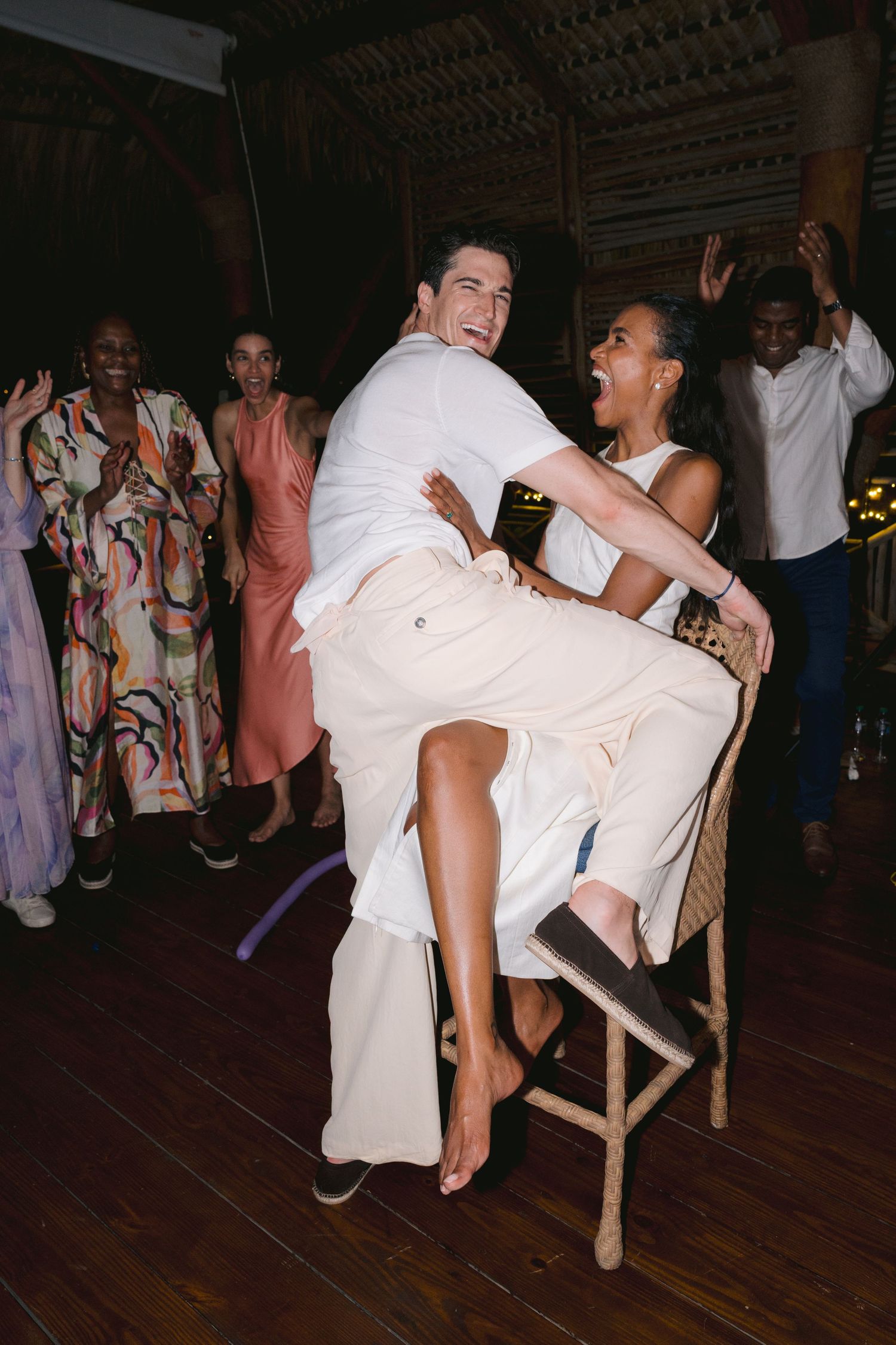 A couple in all-white outfits dances energetically at a celebration while guests in colorful attire cheer in the background.