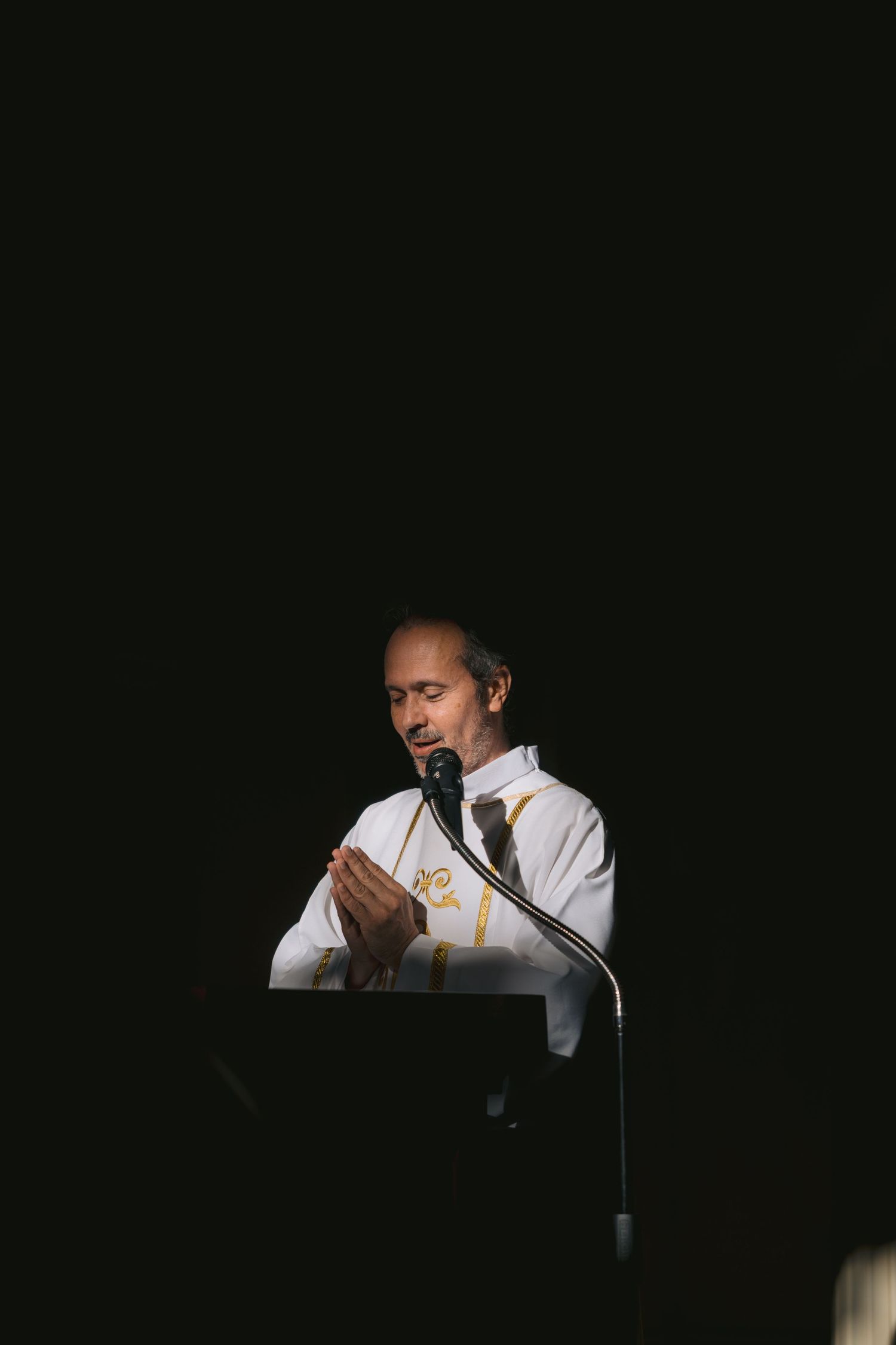 Speaker at podium reads from papers in darkened room with microphone and dramatic lighting.