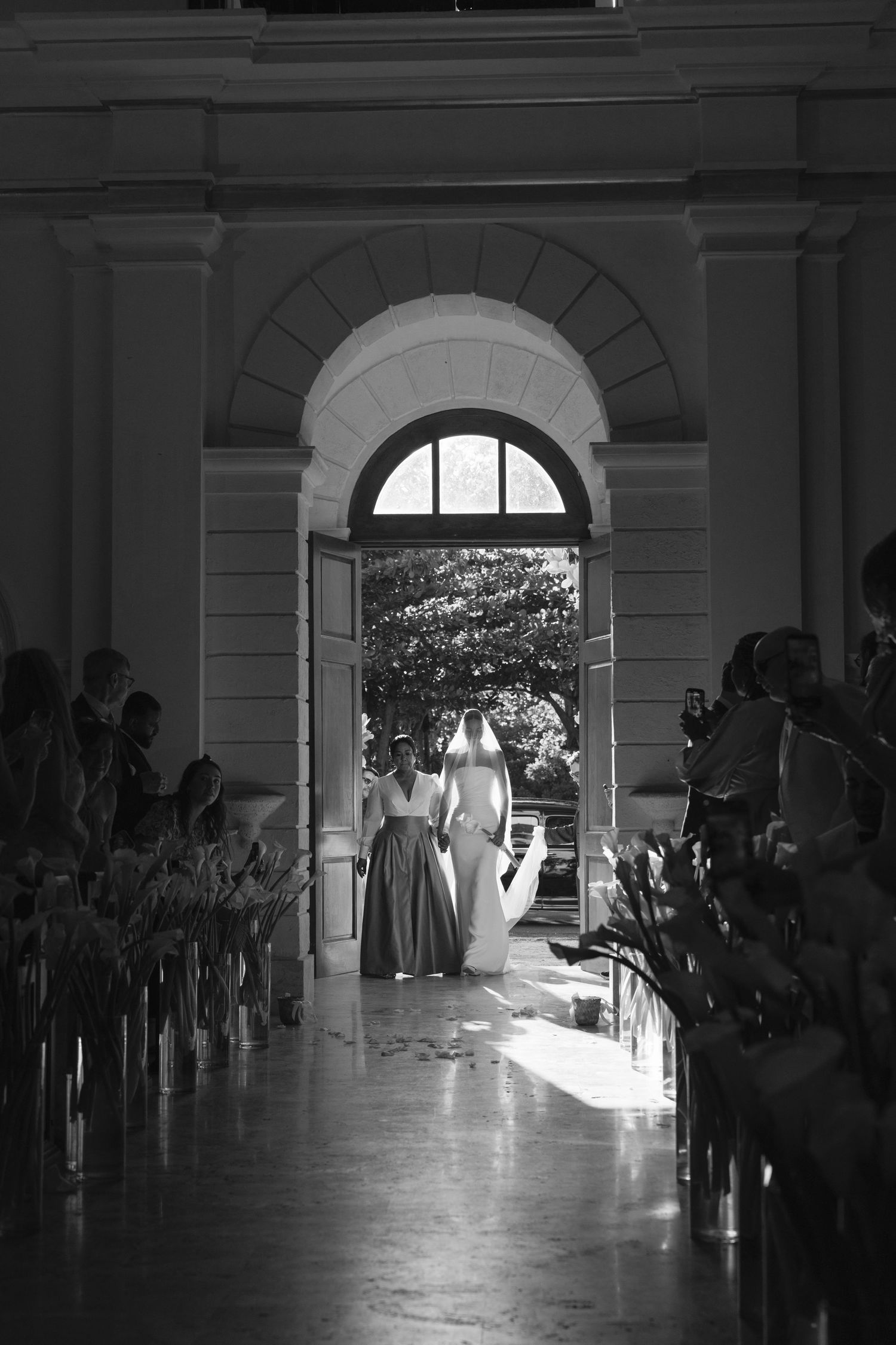 Black and white photograph of guests seated along a corridor with natural light streaming through an arched doorway.