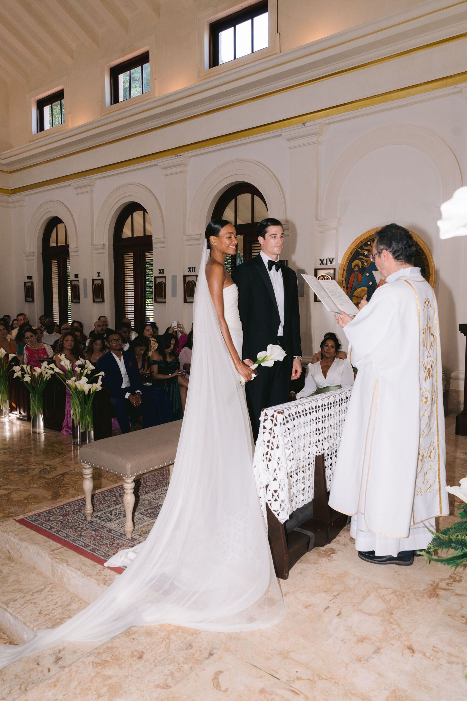 A traditional Catholic wedding ceremony taking place in an elegant marble church with arched windows and floral decorations.