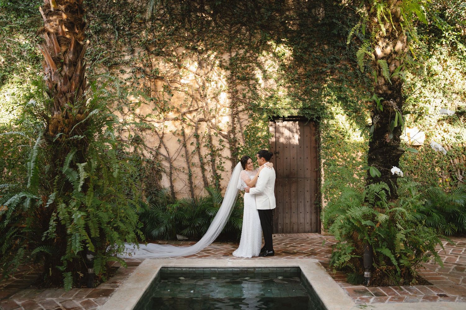 Romantic wedding portrait by a rustic wooden door and reflection pool surrounded by lush greenery.