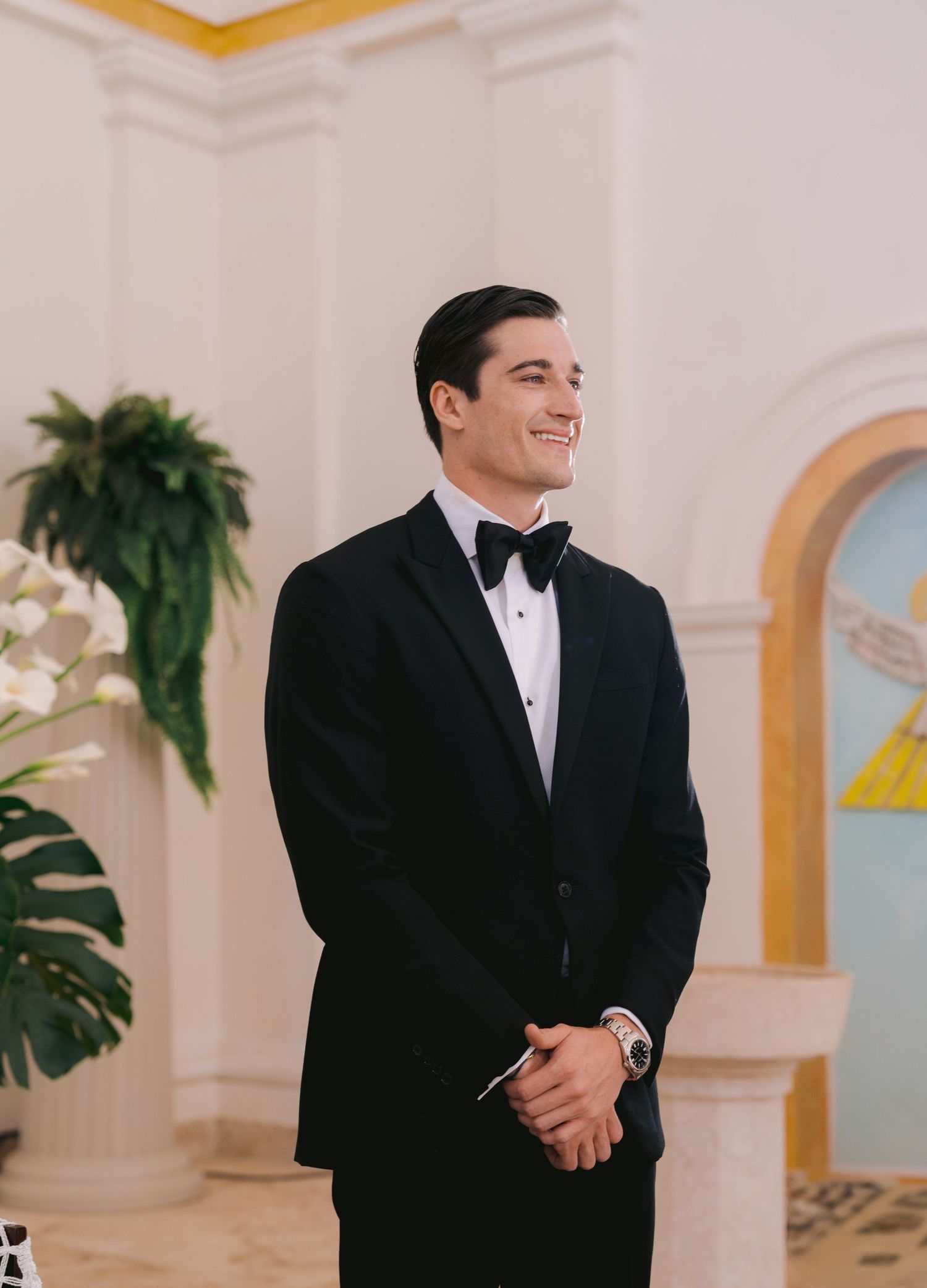 A person in a classic black tuxedo and bowtie smiles warmly while standing in an elegant white room with arched windows.