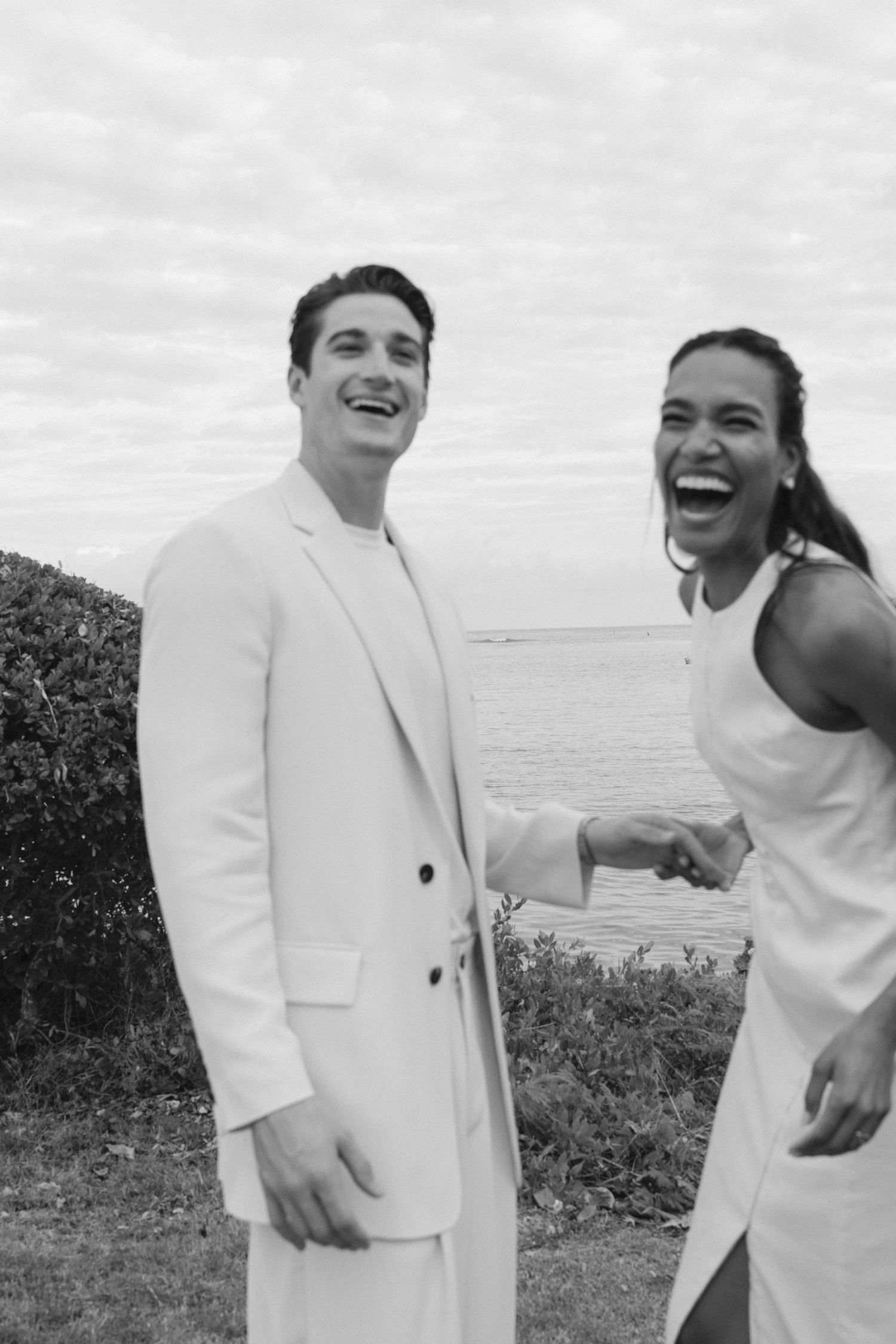 A couple in matching white outfits share genuine laughter outdoors against a cloudy sky beside green foliage.