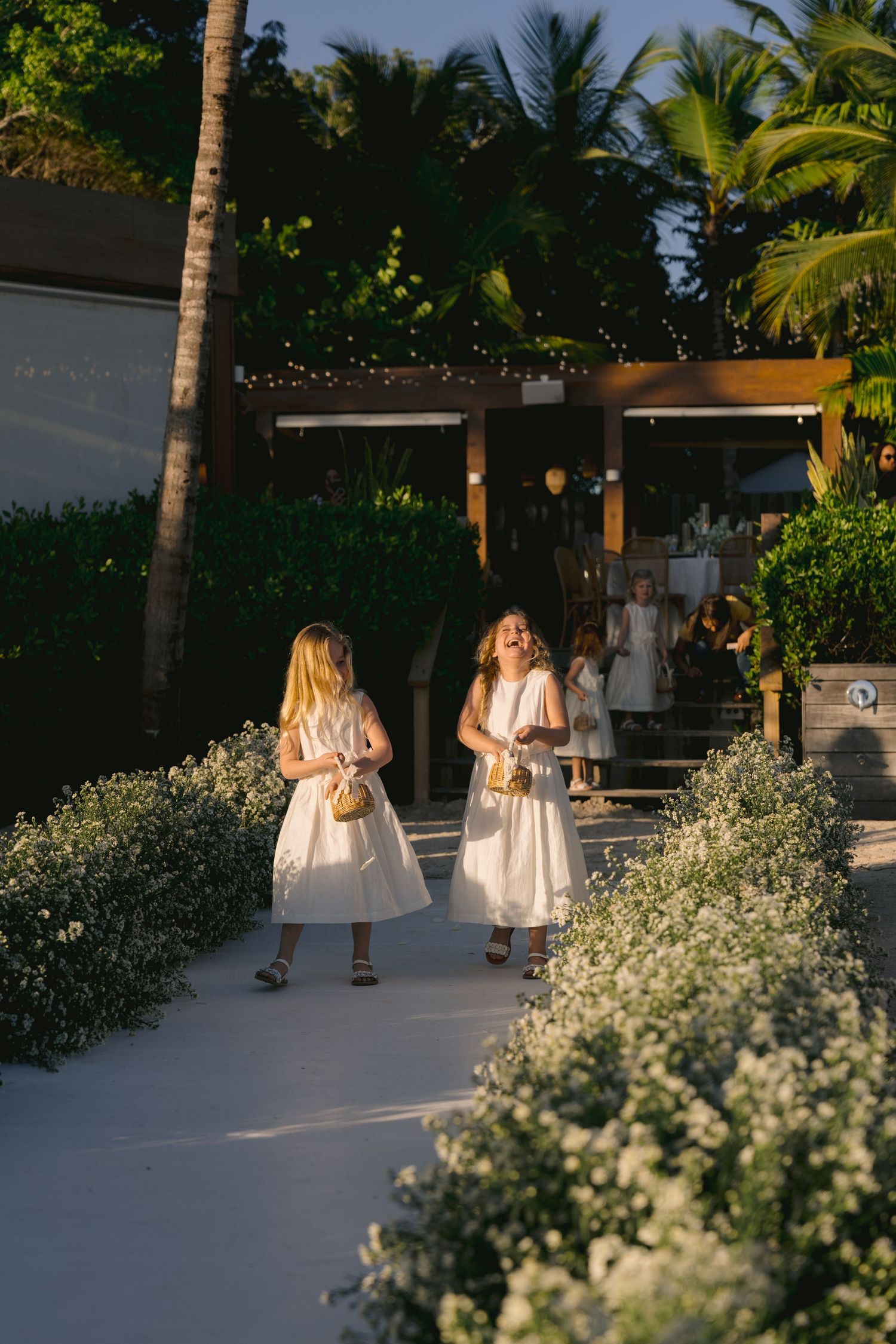 People walking along a tropical resort pathway lined with white flowers and palm trees at sunset.