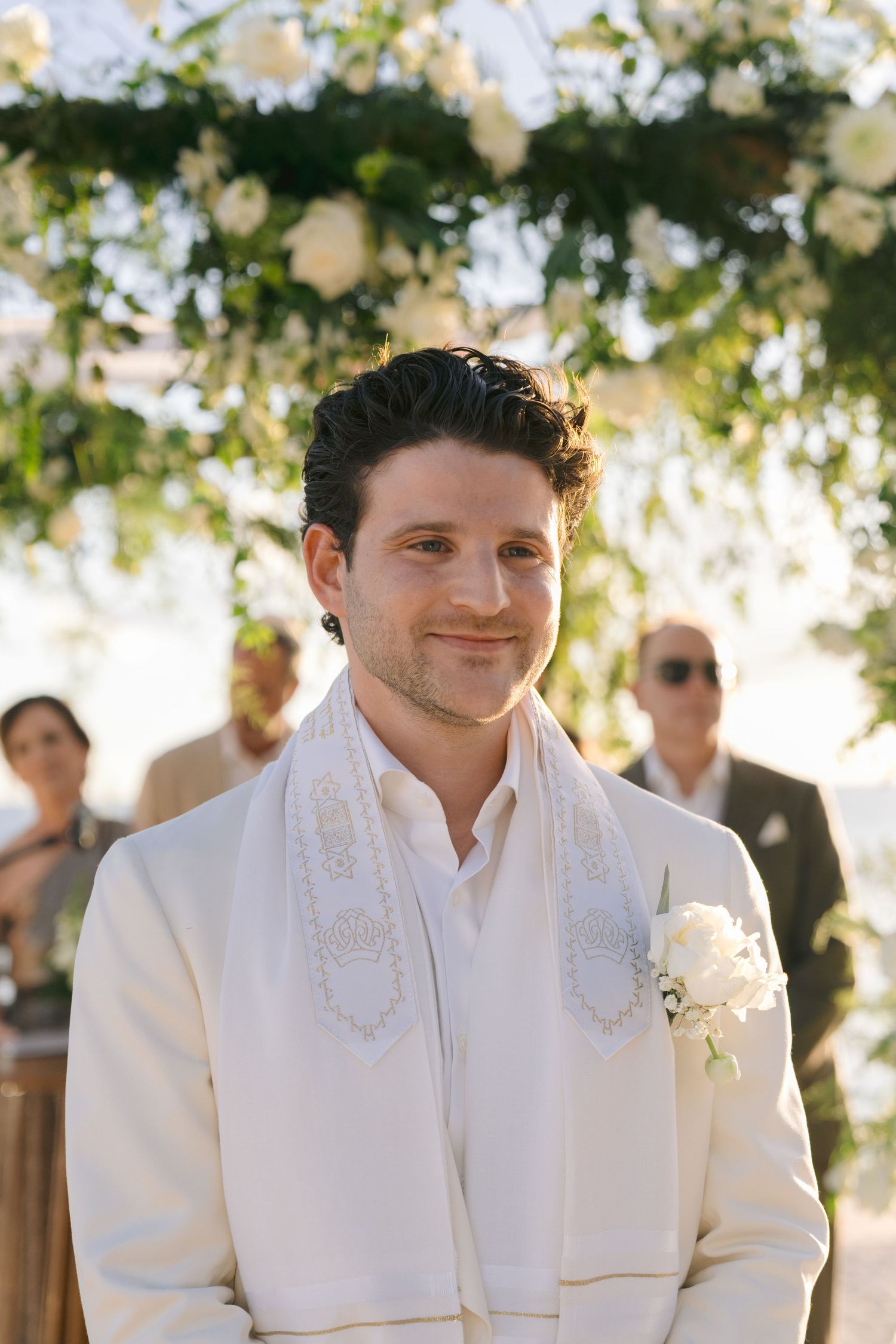 A person in a white collared shirt smiles warmly during an outdoor event with sunlit trees in the background.