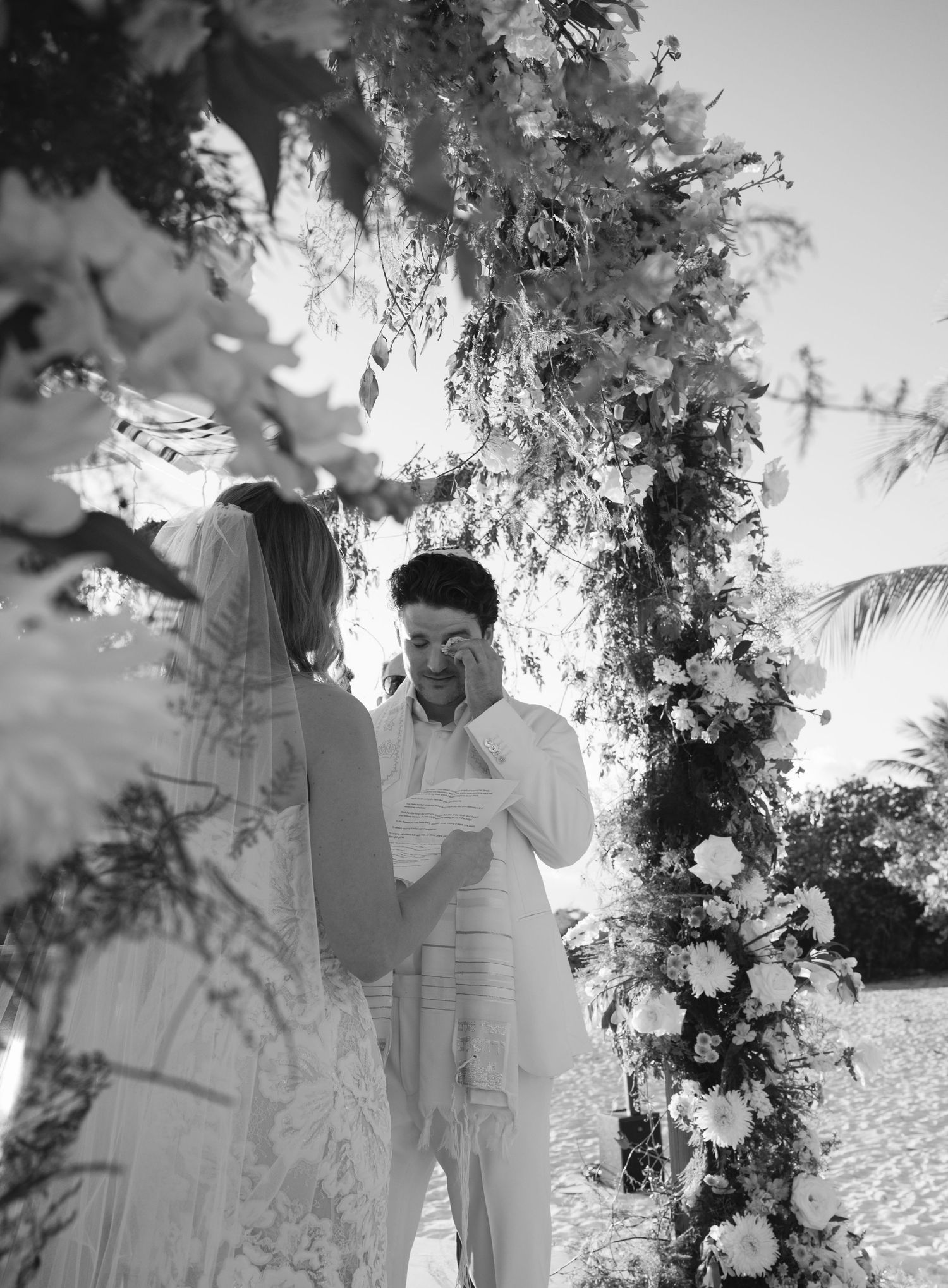 A romantic black and white photo series shows a couple in white attire embracing under floral arches on a beach.