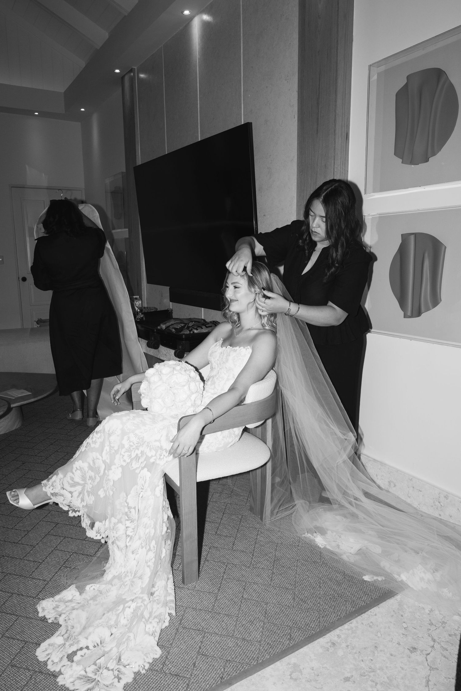 A black and white photo shows a bride in a lace wedding dress having her veil styled in a bridal preparation room.