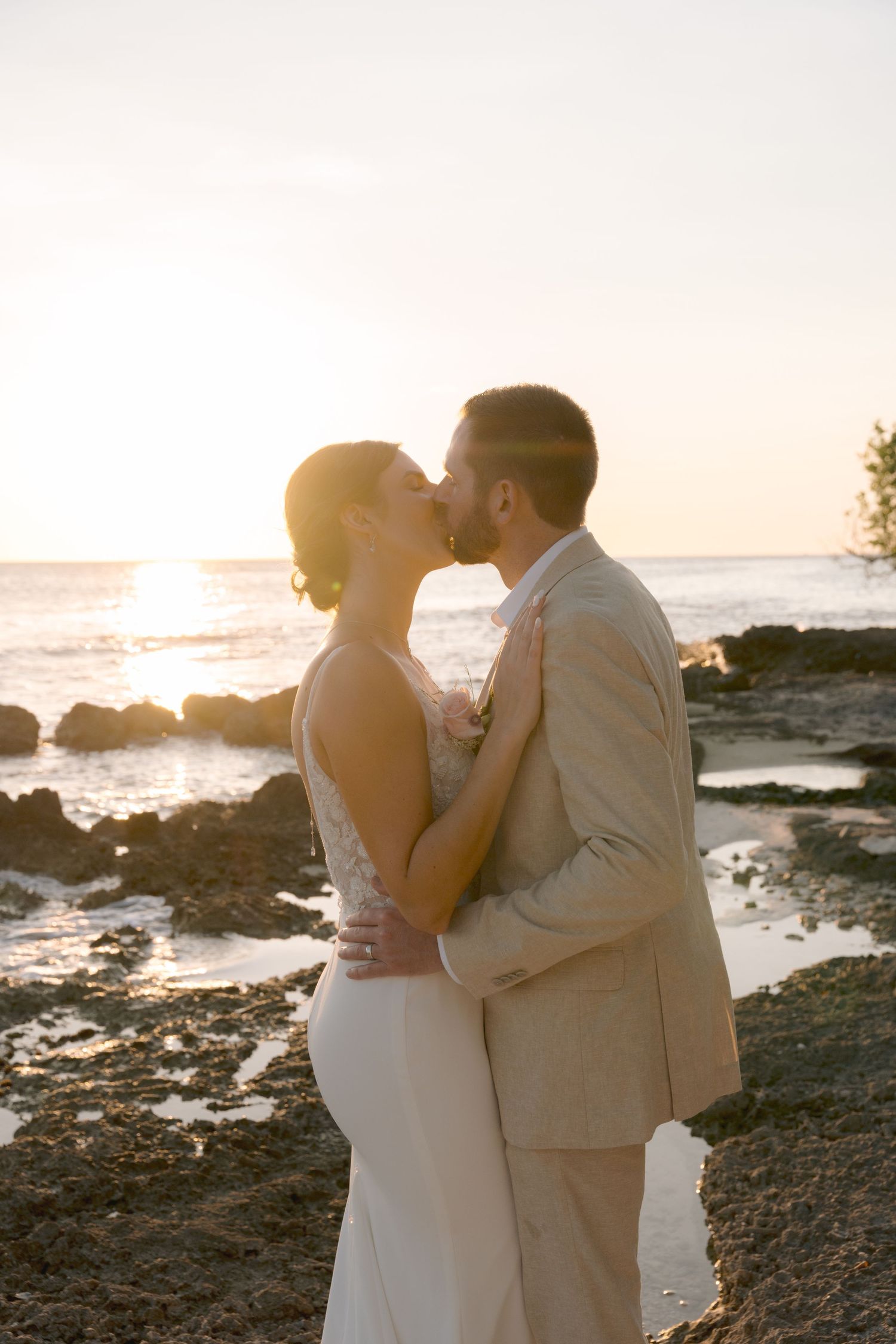 A romantic wedding couple shares a sunset kiss on a rocky beach shoreline.