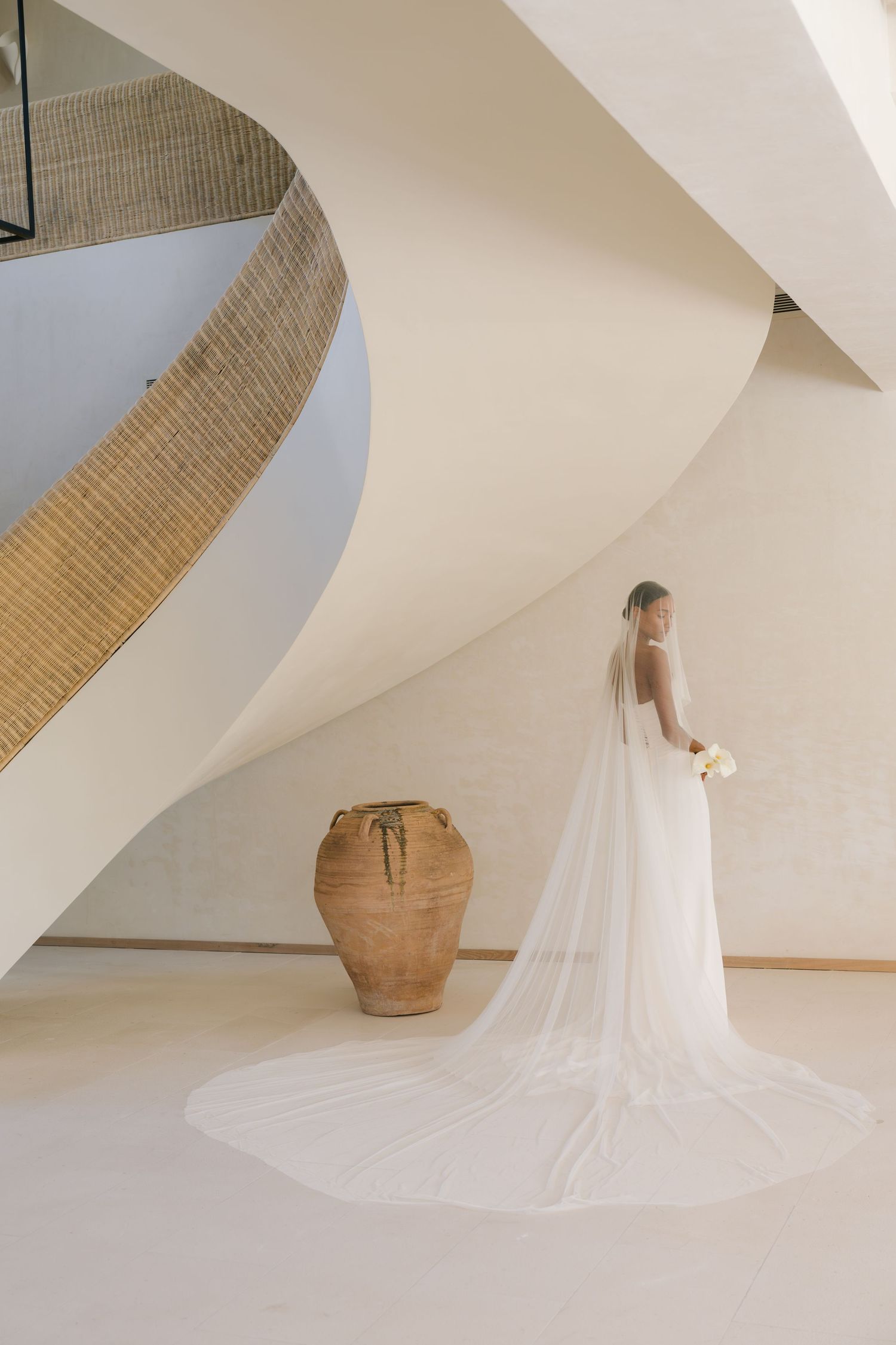 A bride in a flowing white gown with long veil stands beside a large terracotta pot near a curved staircase.